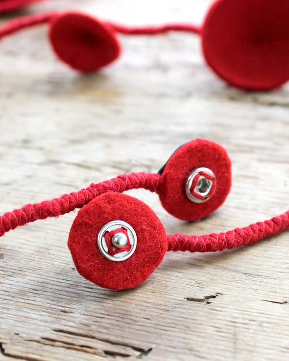 Showing the clasp red braided cord with round felt-like button charms and silver metal rivets, photographed on a rustic light wood surface.