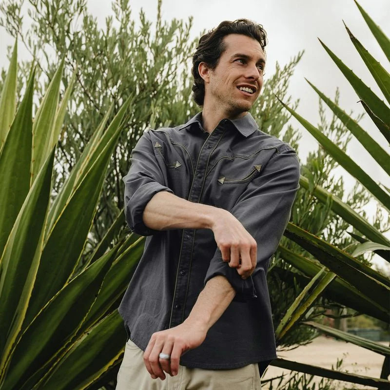 A man wearing a dark charcoal gray button-down shirt with gold-toned embroidery on the chest, standing outdoors in front of green plants.