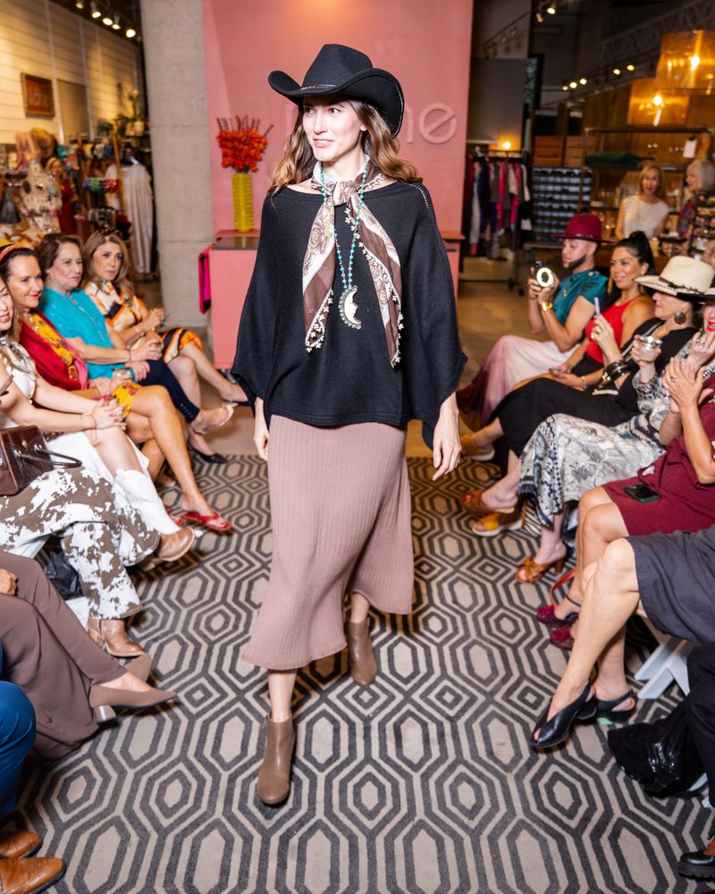 model wearing a black cowboy hat, black poncho, knit taupe skirt, brown scarf, long turquoise beaded moon necklace and boots walking down a runway
