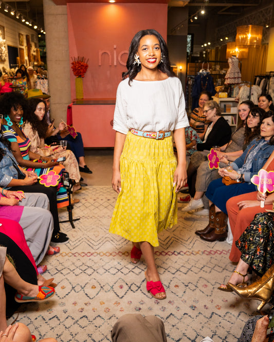 Woman in a white top and yellow skirt standing in front of an audience in a store setting.