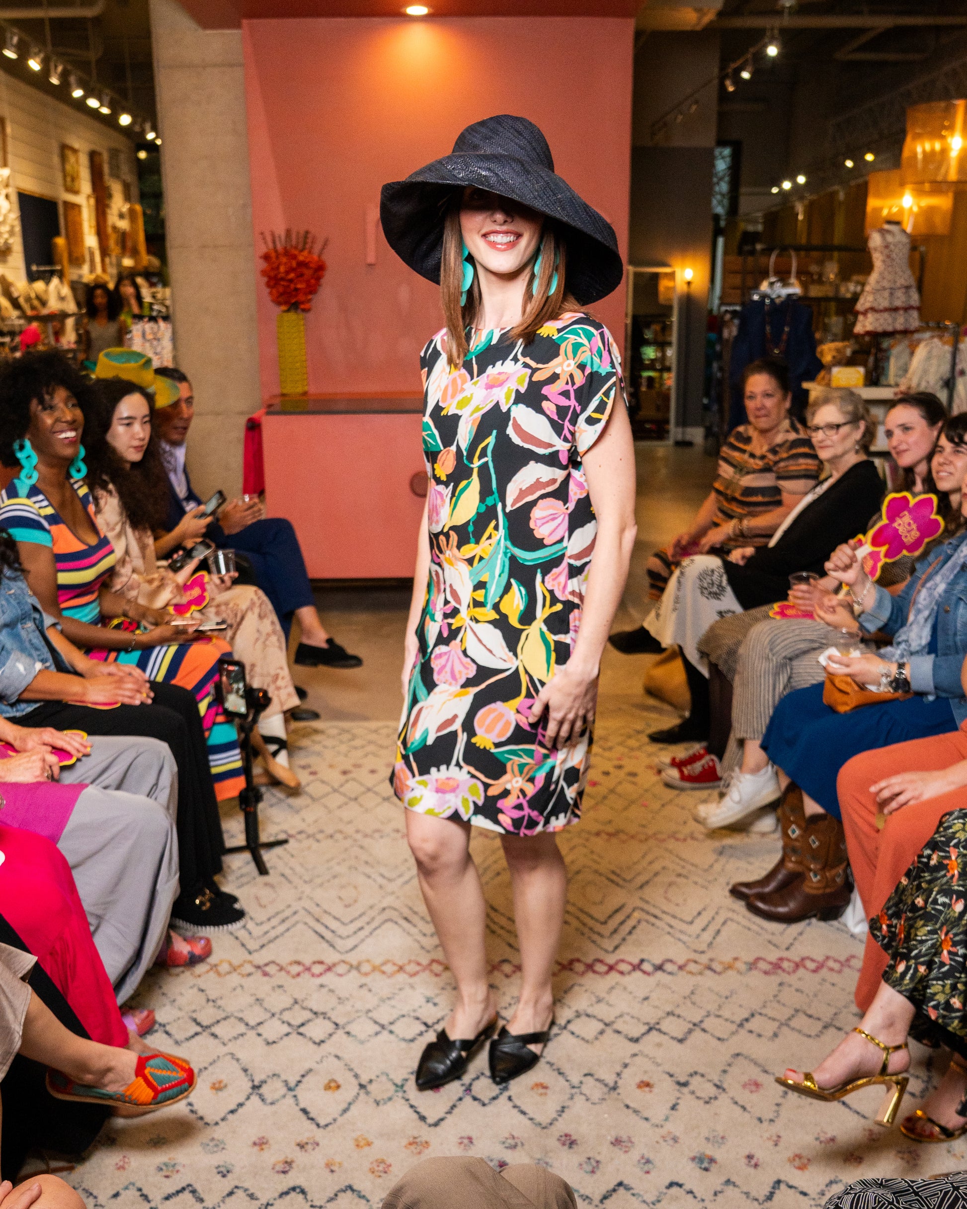 Woman in a colorful dress and large hat at a fashion event with an audience.