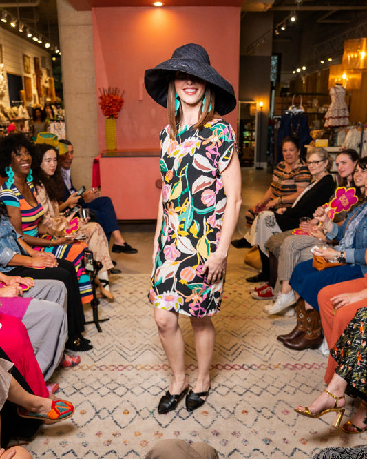 Woman in a colorful dress and large hat at a fashion event with an audience.