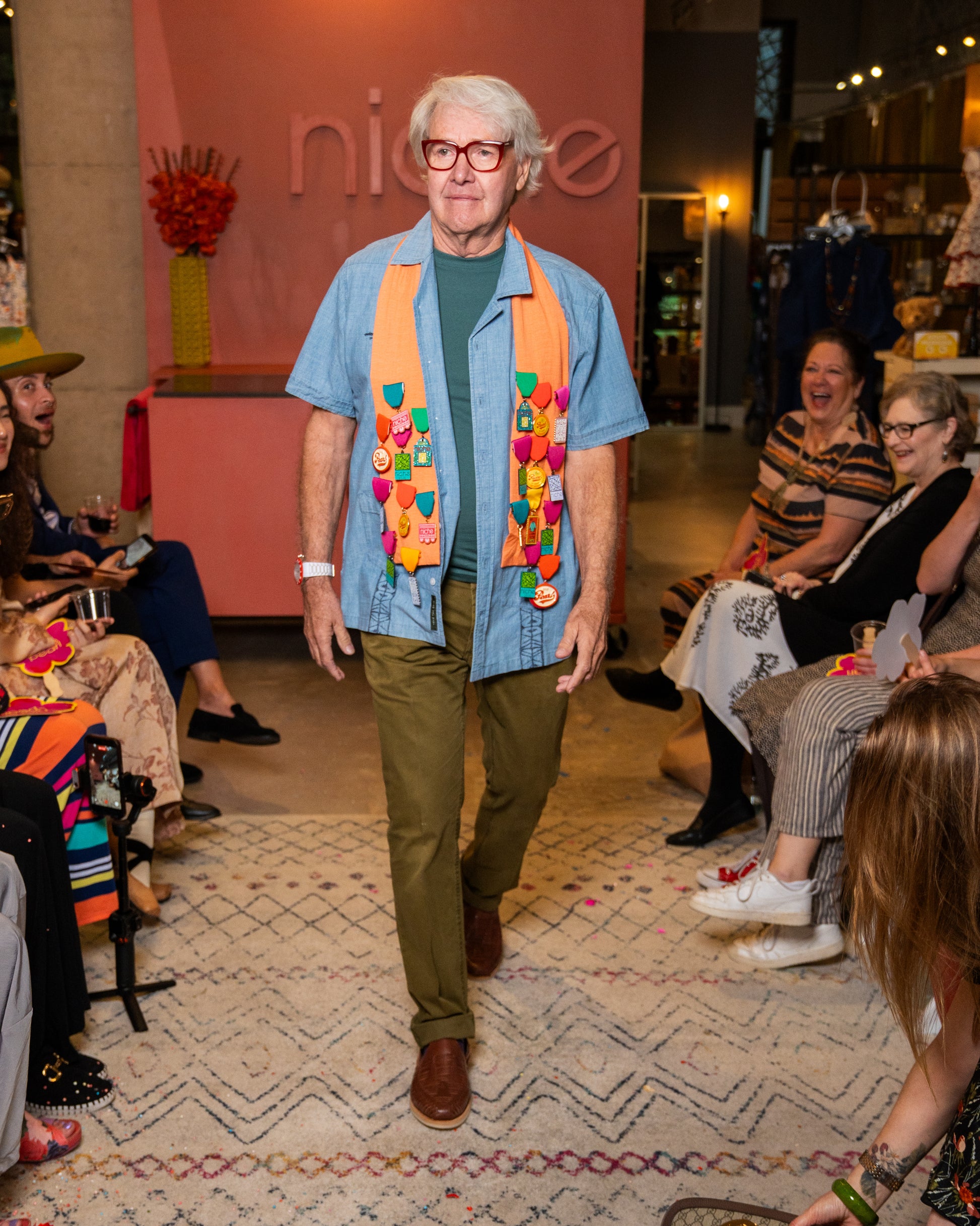 Man wearing a colorful sash, blue shirt and green pants while walking down a runway in a store setting with people around