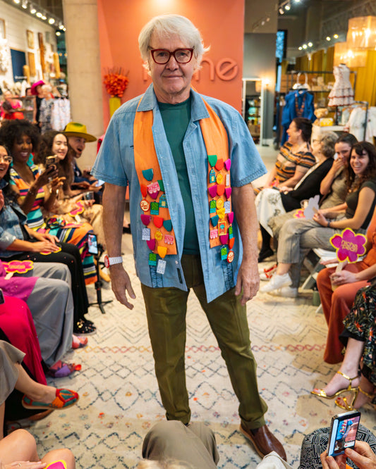 Man wearing a colorful sash, blue shirt and green pants while walking down a runway in a store setting with people around