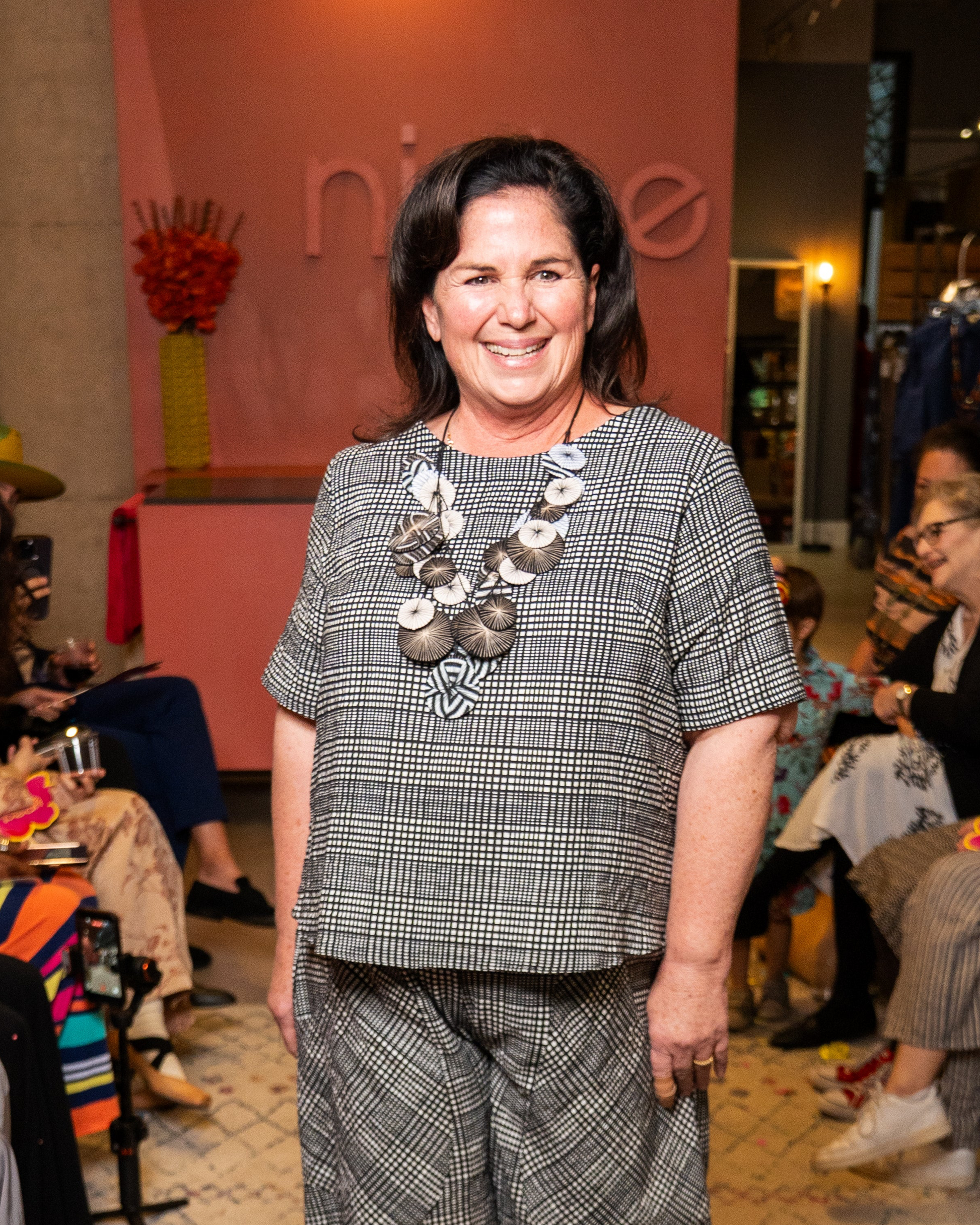 Woman in a checkered outfit with a necklace standing in a social setting.