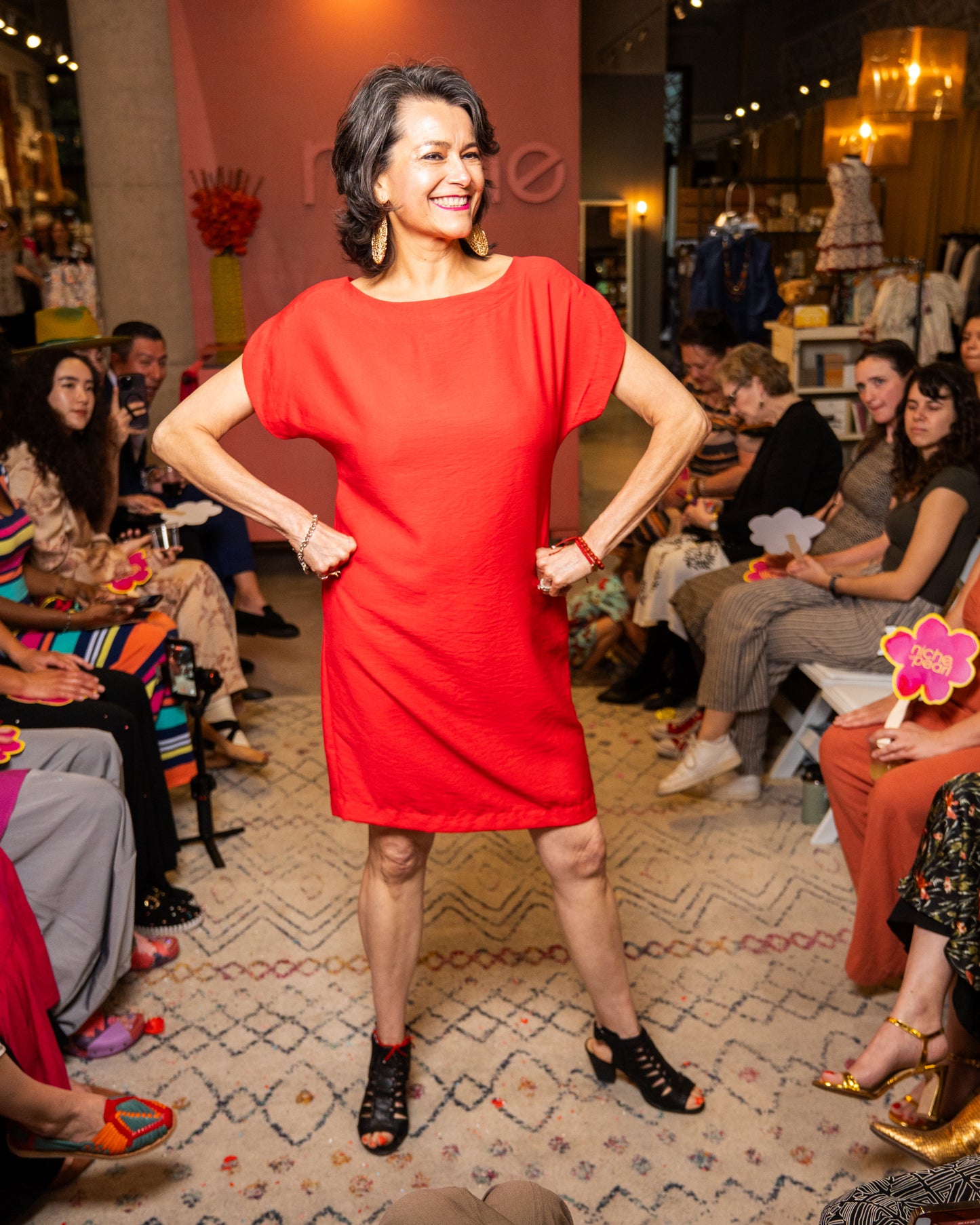 Woman in a red dress posing at a fashion event with an audience in the background.