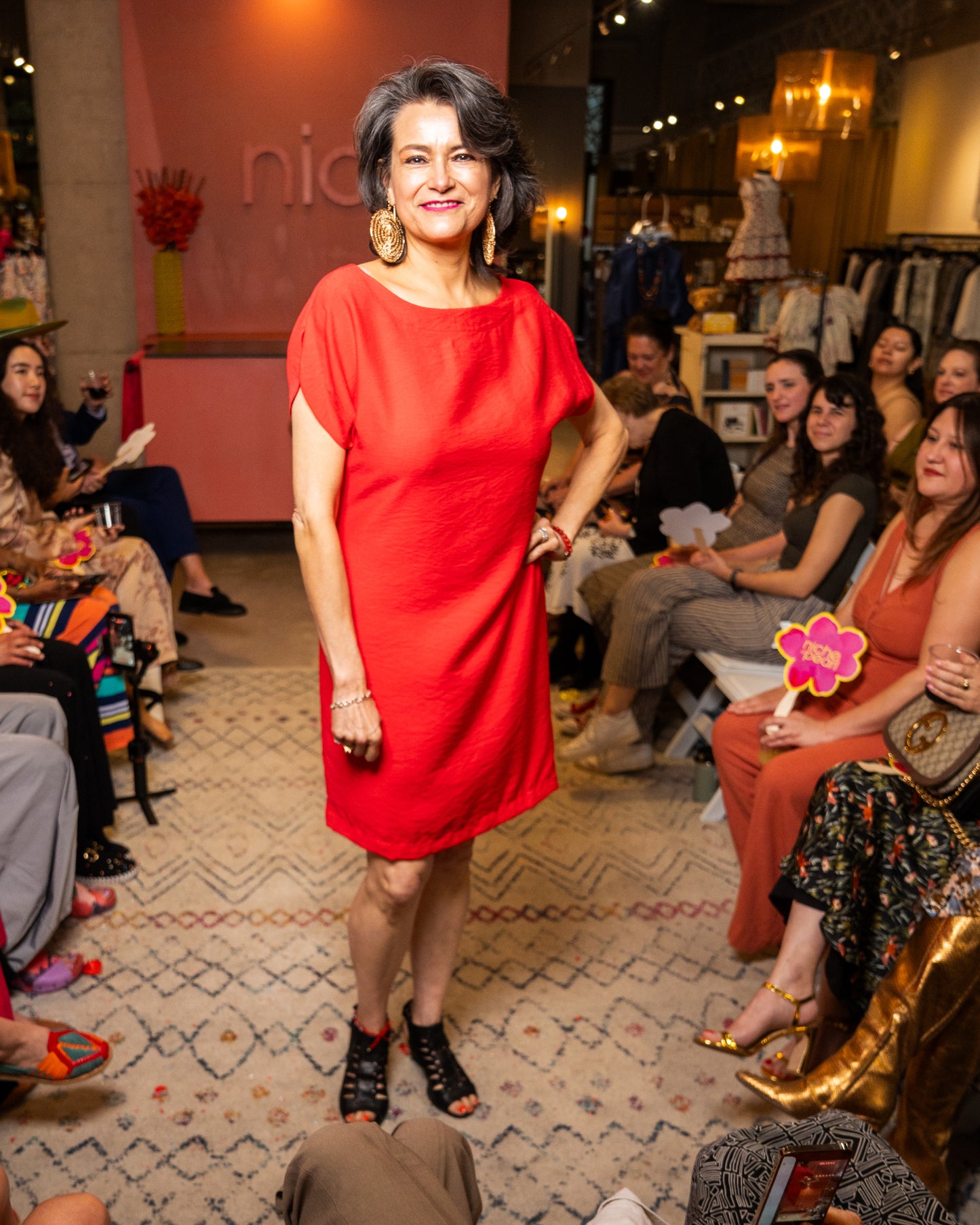 Woman in a red dress standing on a catwalk with an audience in a fashion show setting.