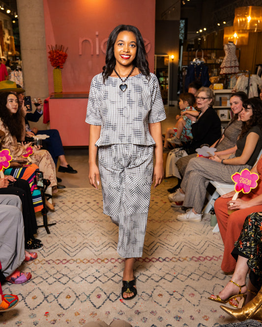 Woman wearing a black and white print top and pants walking on a catwalk at a fashion event with an audience watching.
