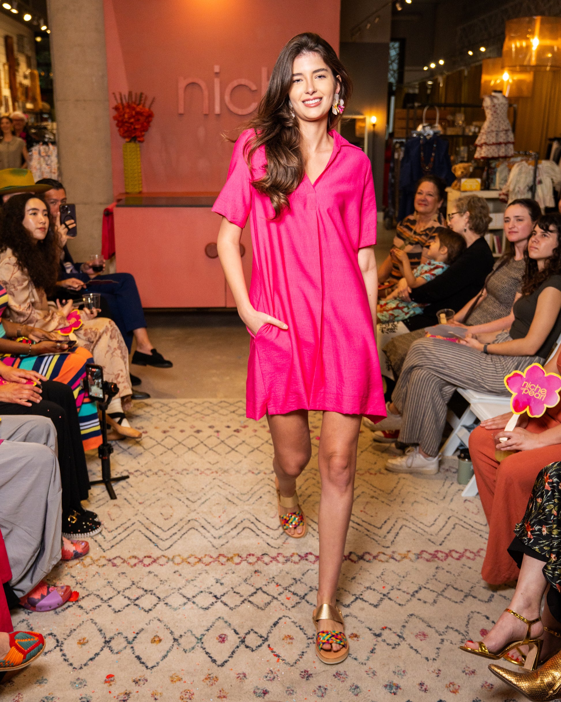 Woman in a bright pink dress walking on a catwalk with an audience watching.