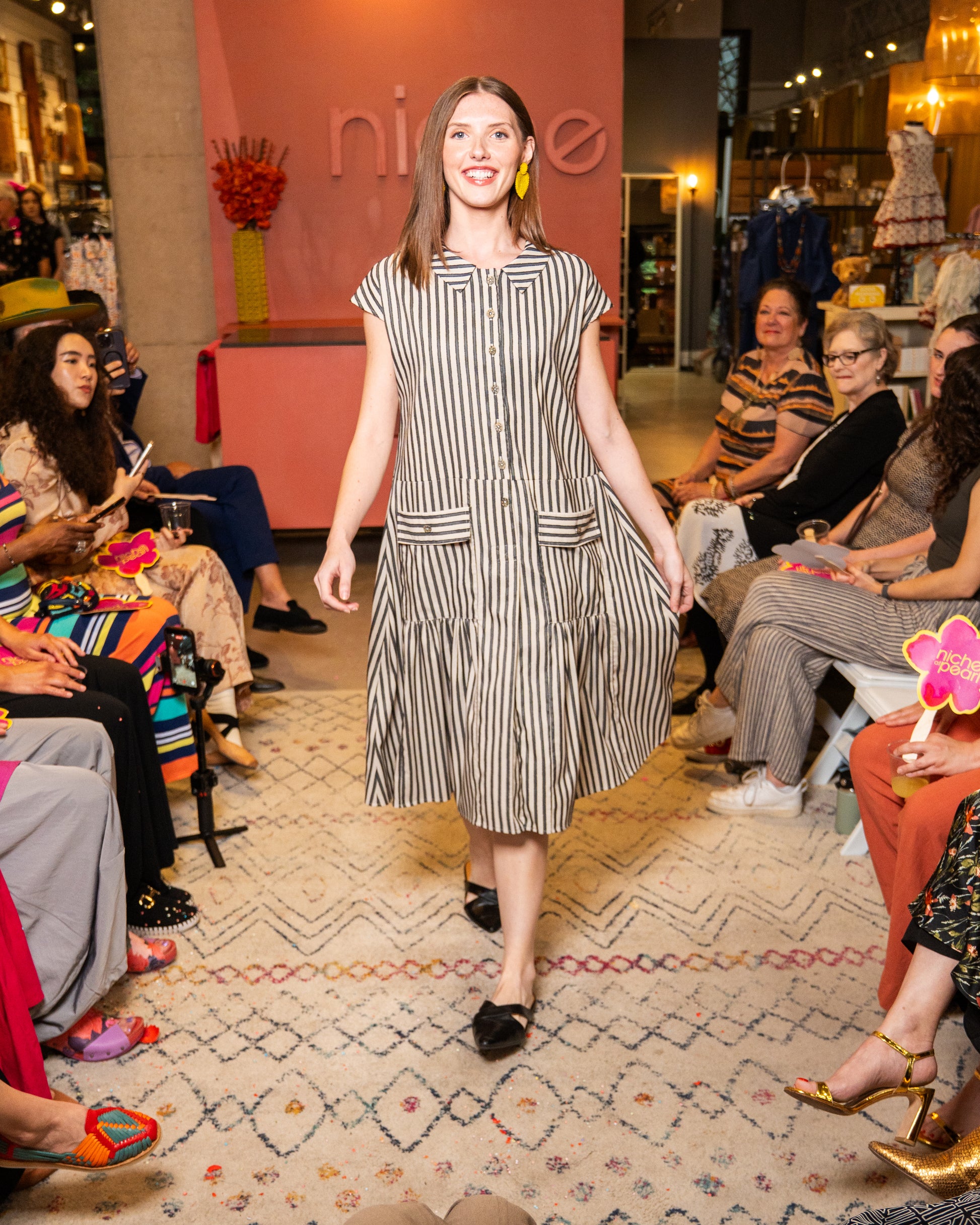 Woman walking on a catwalk wearing a striped black and white dress at a fashion event with an audience watching.