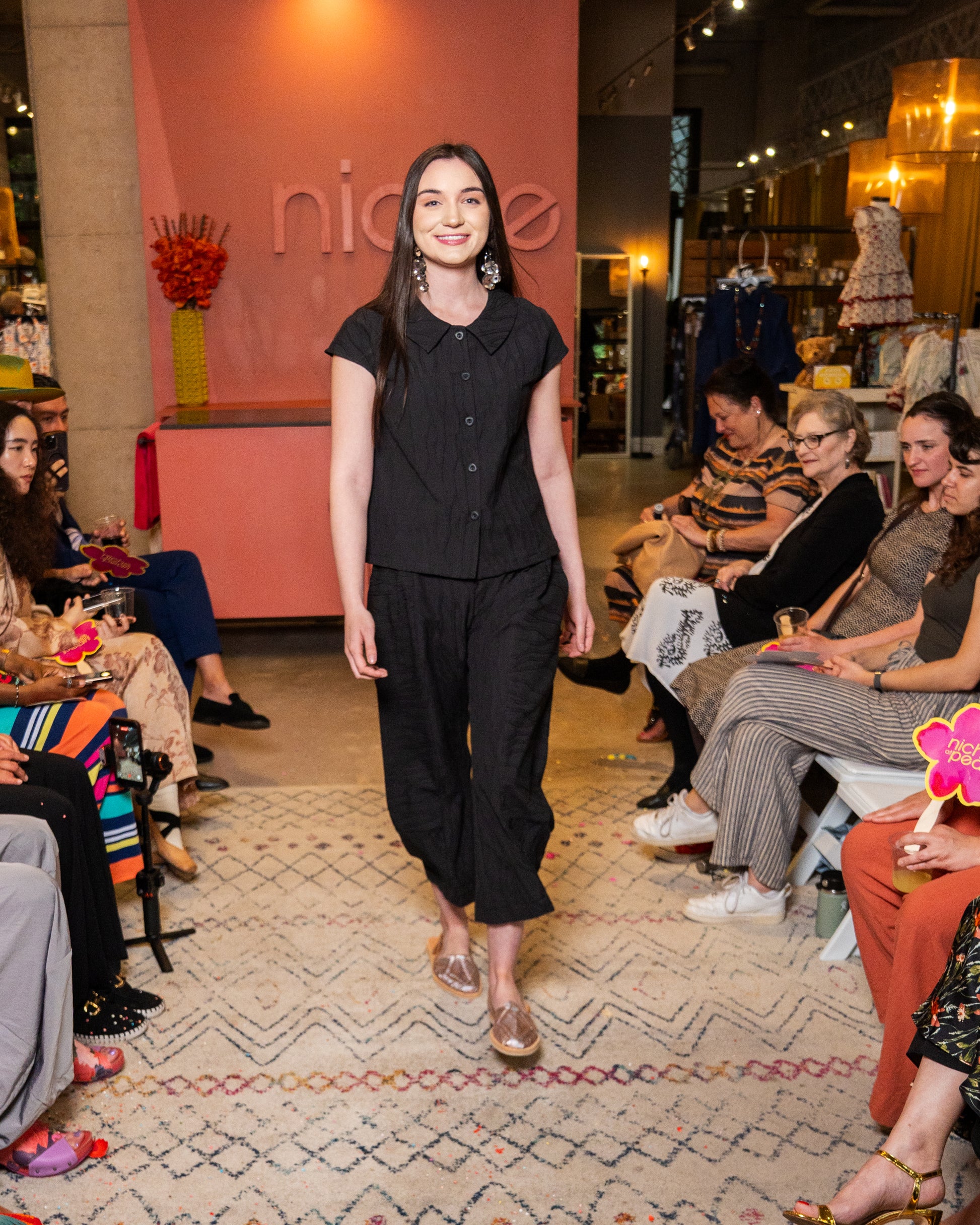 Woman in black top and pant walking on a runway with an audience watching.