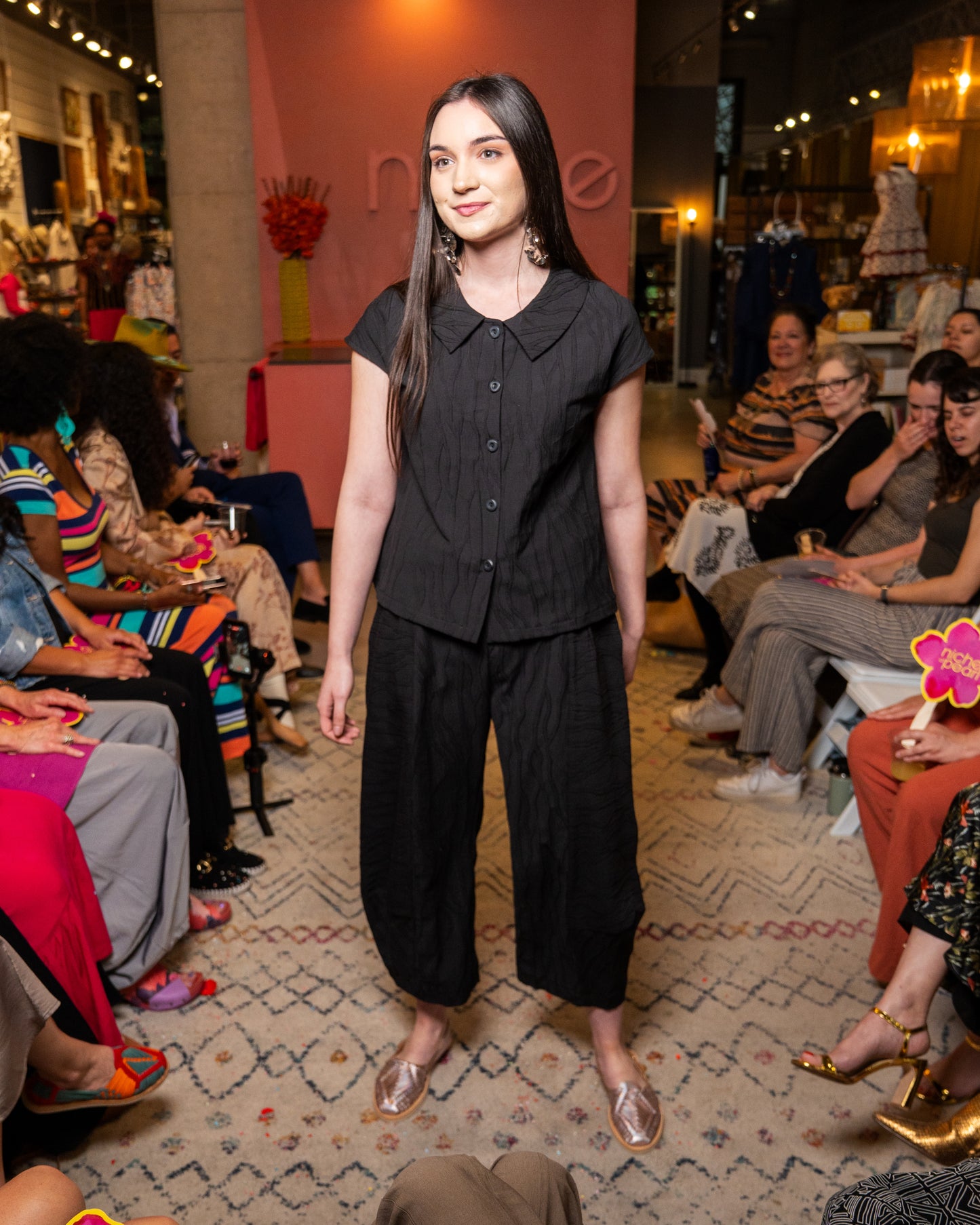 Woman in black top and pant walking on a runway with an audience watching.