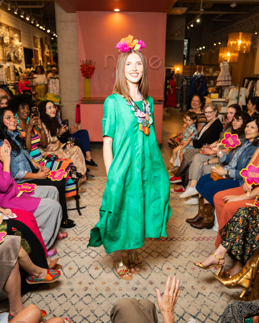 Model in a green dress with colorful accessories walking on a runway in front of an audience.