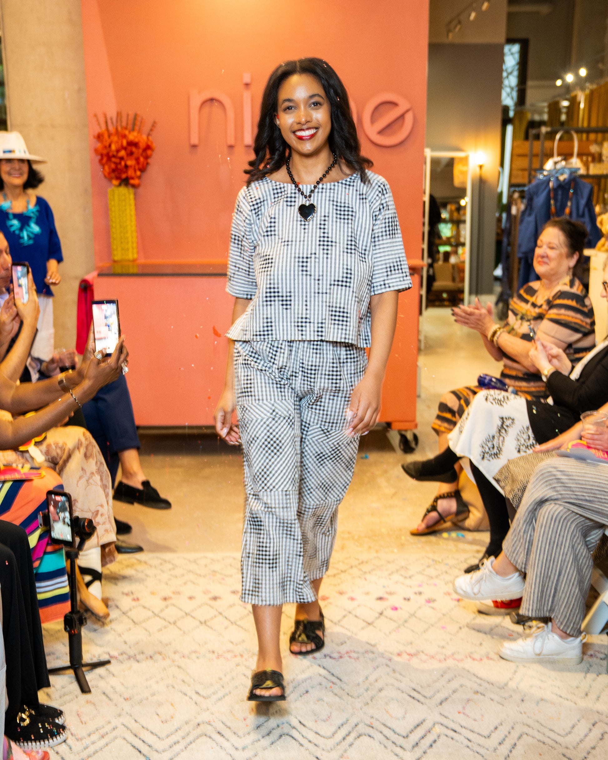 Woman wearing a black and white print top and pants walking on a catwalk at a fashion event with an audience watching.
