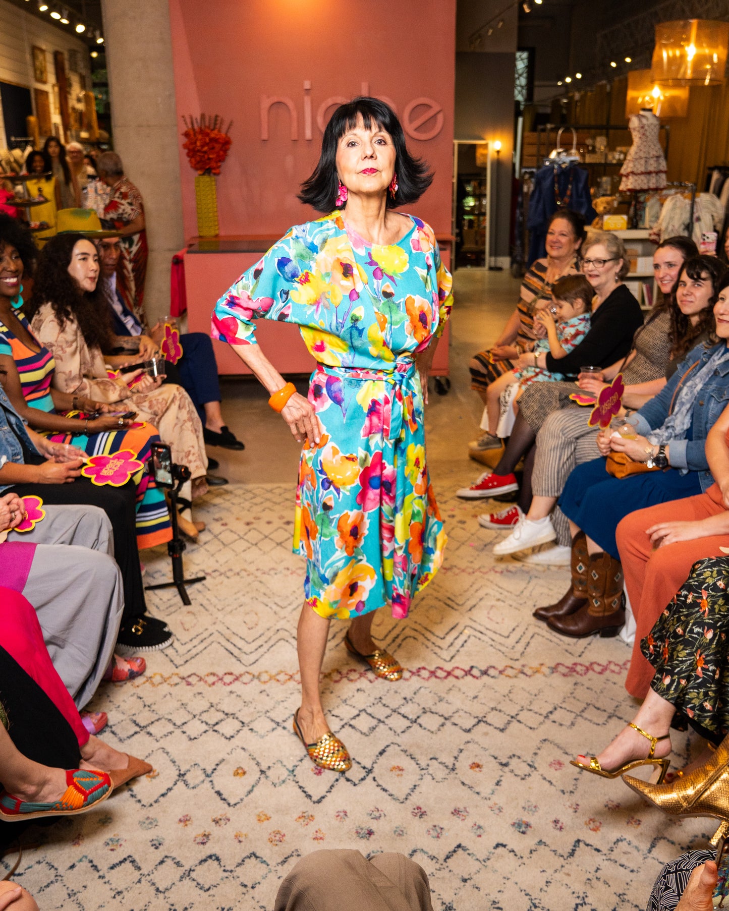 Woman in a colorful dress walking on a catwalk with an audience watching.