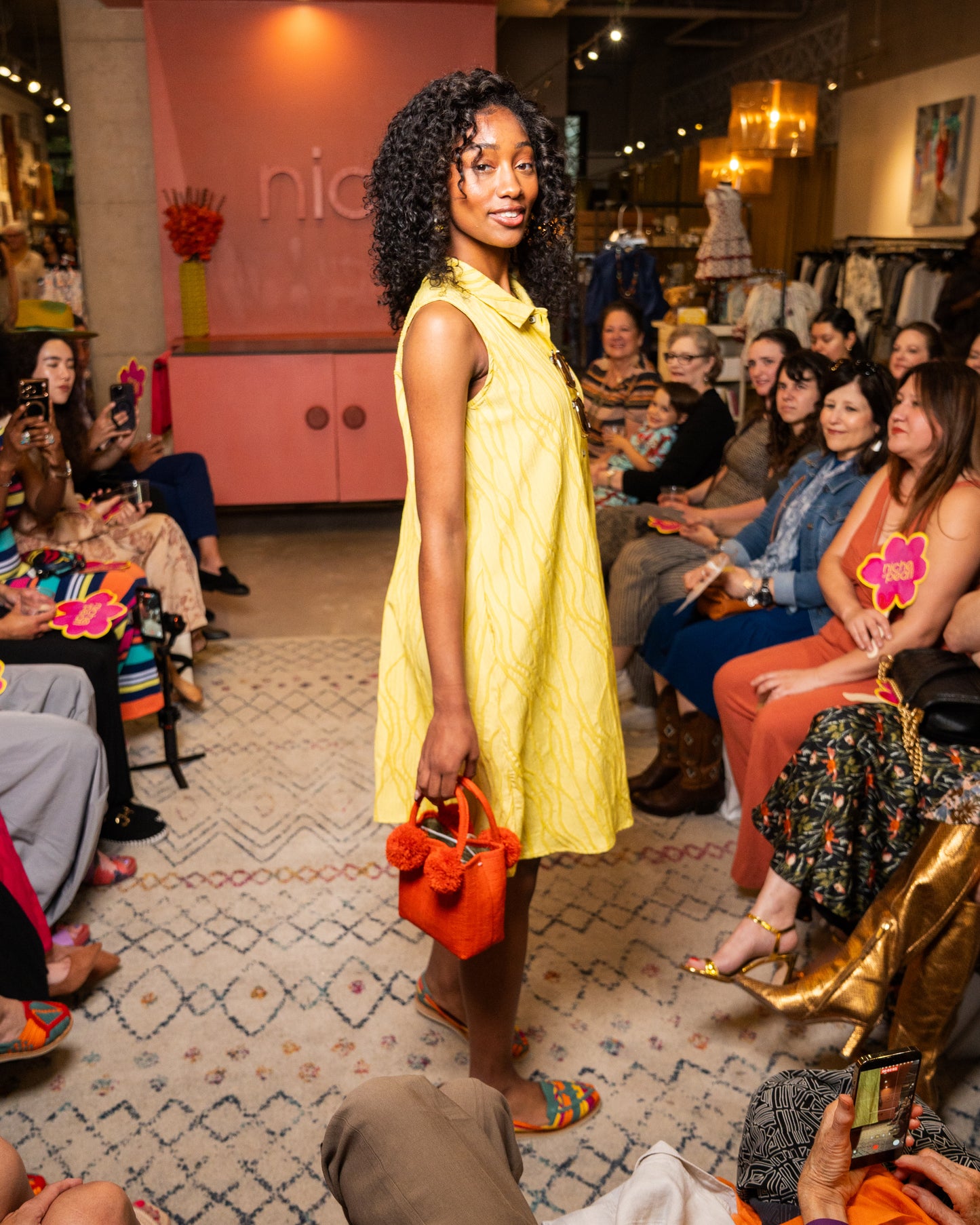 Woman in yellow dress holding an orange handbag at a fashion event with audience.