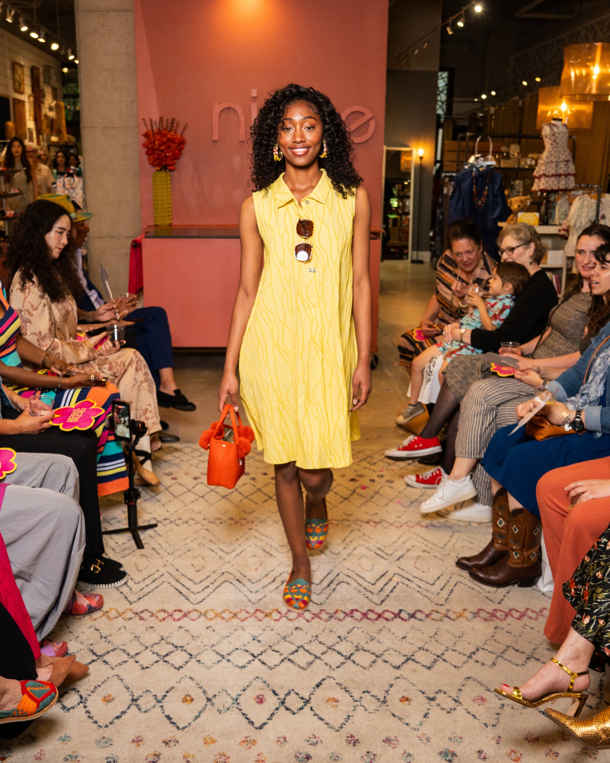 Woman in a yellow dress holding an orange handbag at a fashion event.
