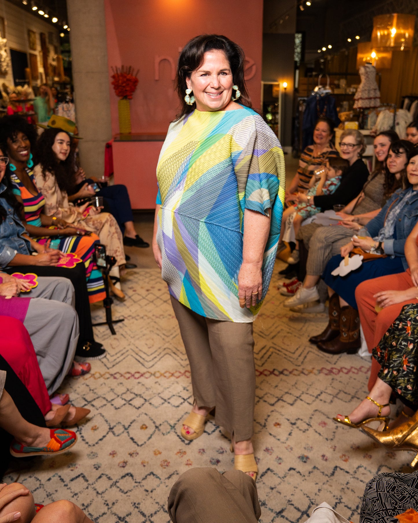 Woman in a colorful top and taupe pants walking down a runway.
