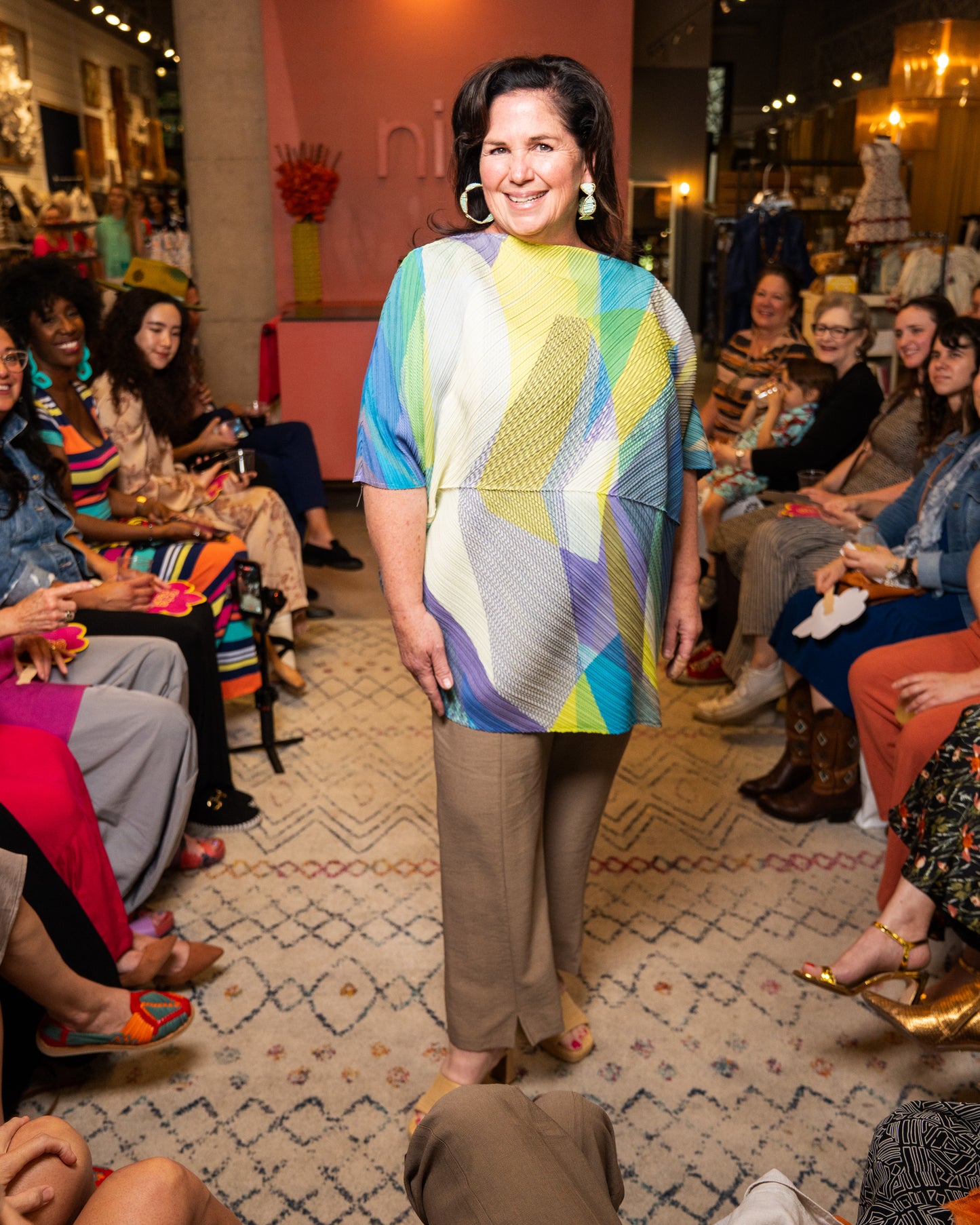 Woman in a colorful top and taupe pants walking down a runway.