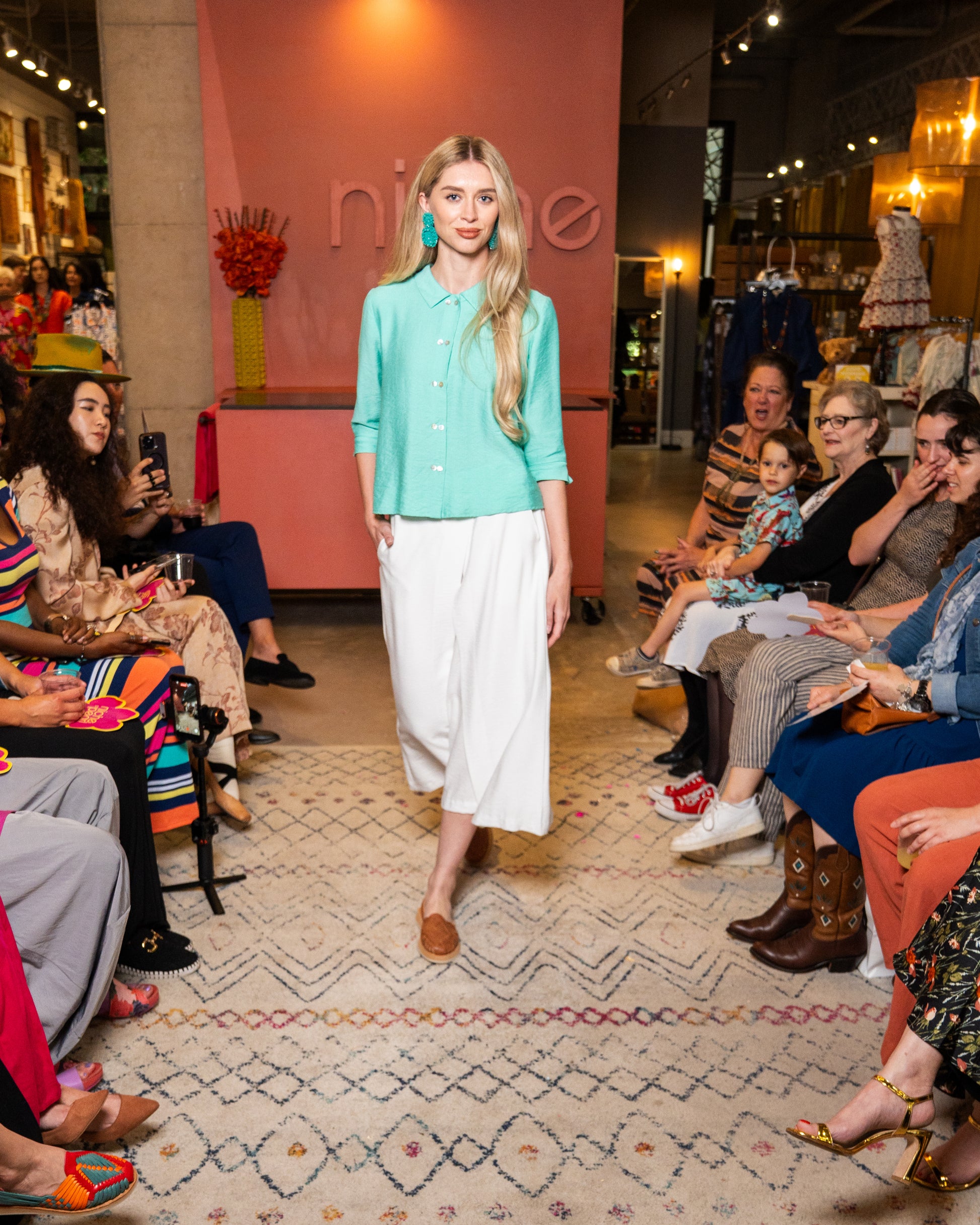 Woman in a mint top and white pants walking on a runway with an audience watching.