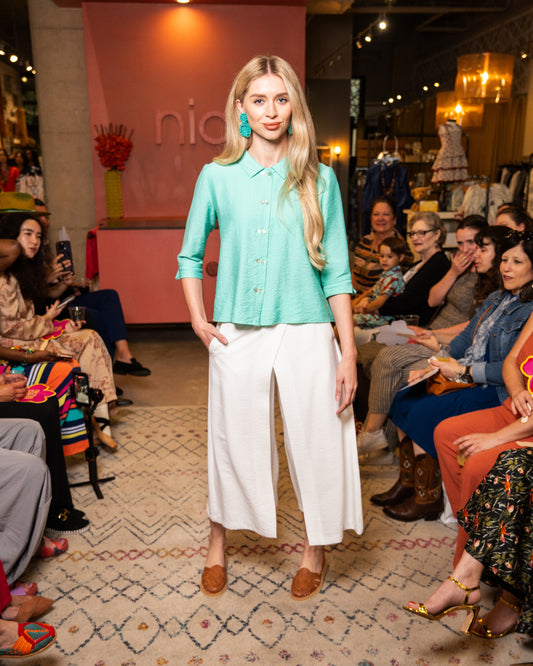 Model wearing a teal blouse and white pants at a fashion event with an audience.