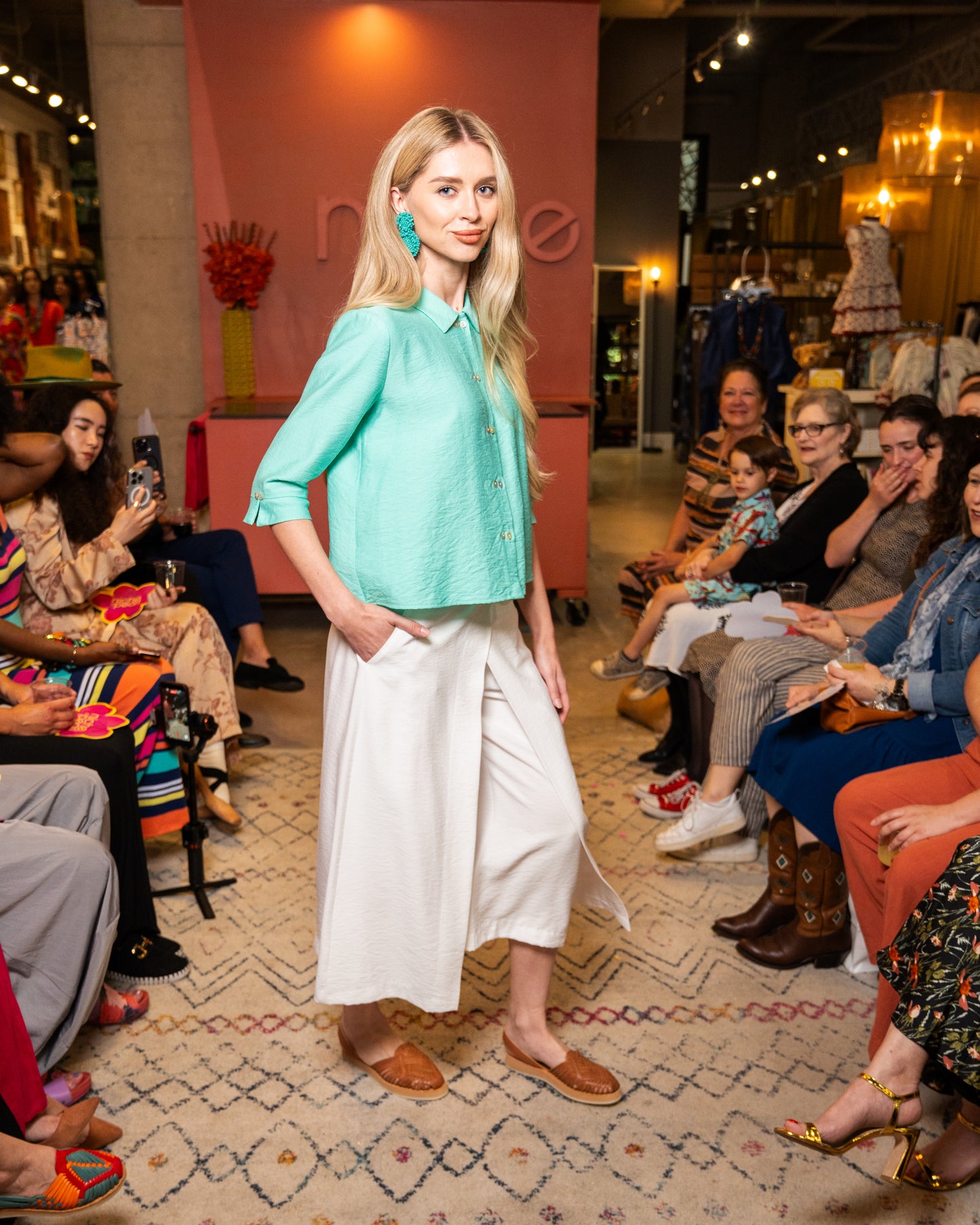 Woman in a teal shirt and white pants standing in front of an audience at a fashion event.