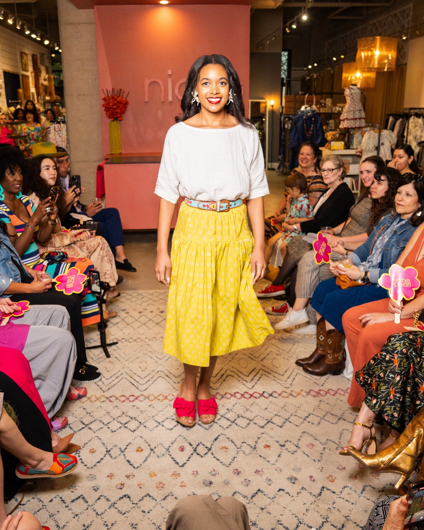 Woman in a yellow skirt and white top standing on a catwalk with an audience in a fashion show setting.