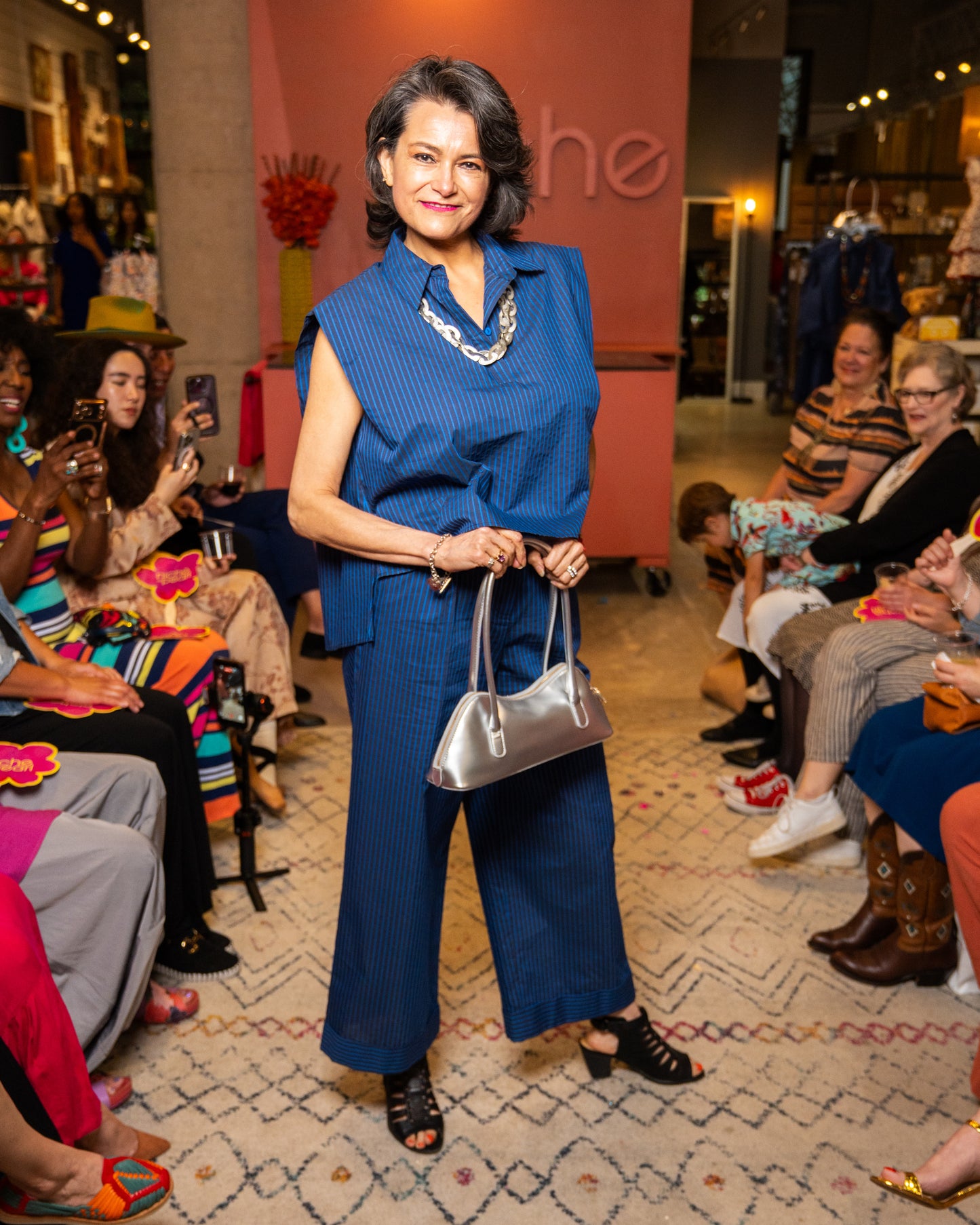 Woman in a blue top and pant and silver necklace and purse walking down a runway at a fashion event