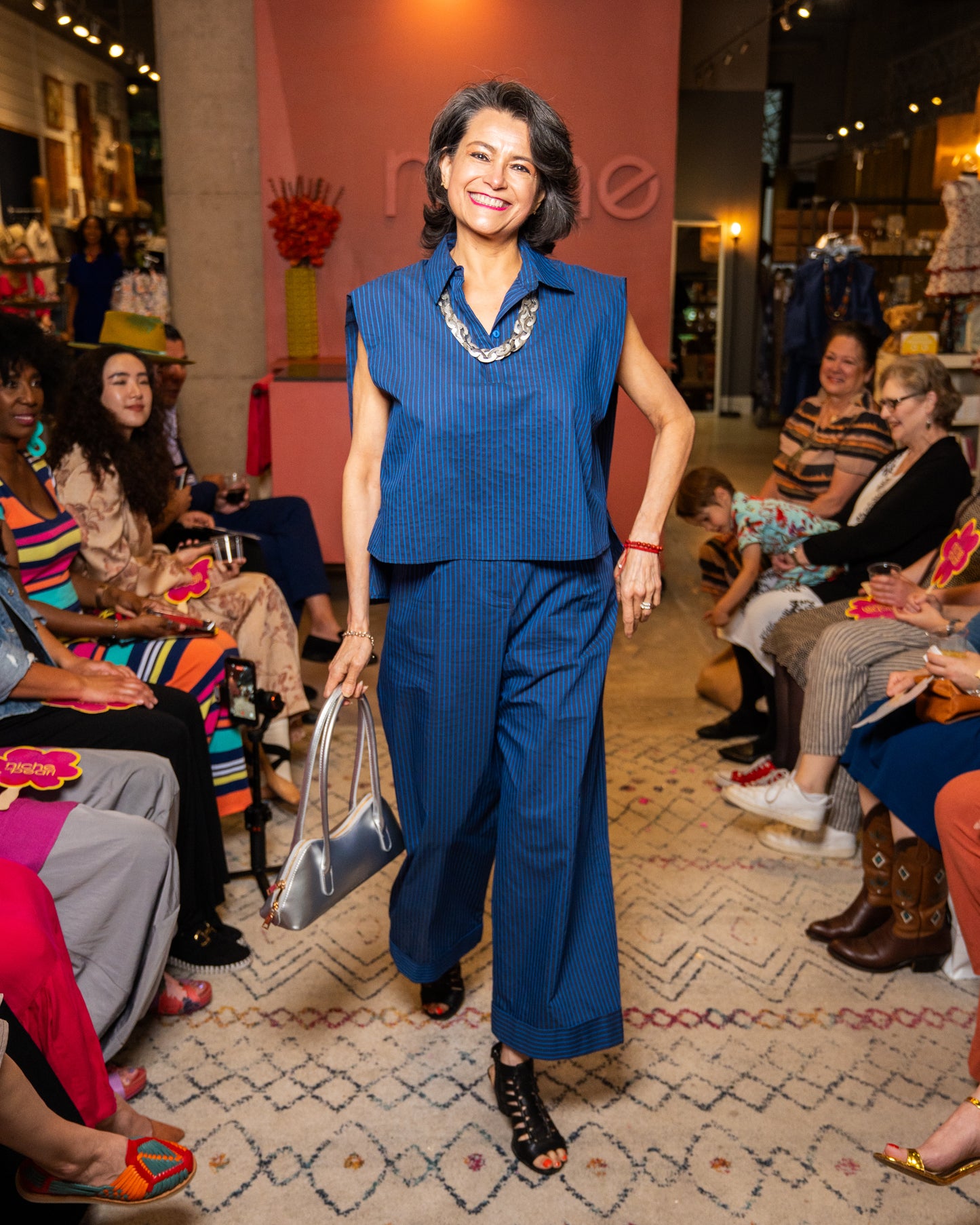 Woman in a blue top and pant and silver necklace and purse walking down a runway at a fashion event