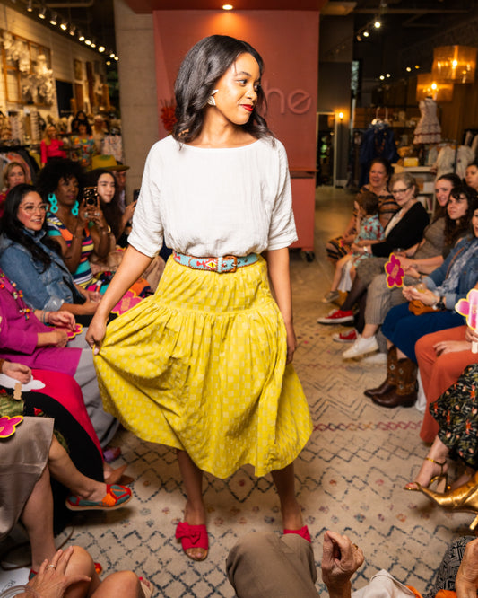 Woman in a yellow skirt and white top walking down a runway with an audience on either side.