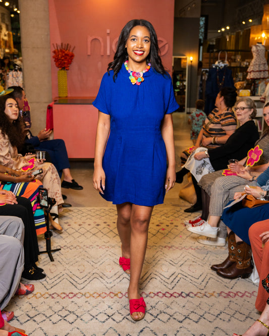 Woman in a blue dress with colorful sandals walking on a catwalk at a fashion event.