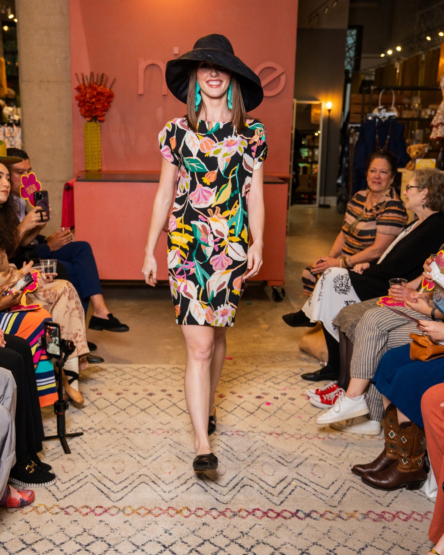 Model walking on a fashion show runway wearing a colorful dress and black hat.