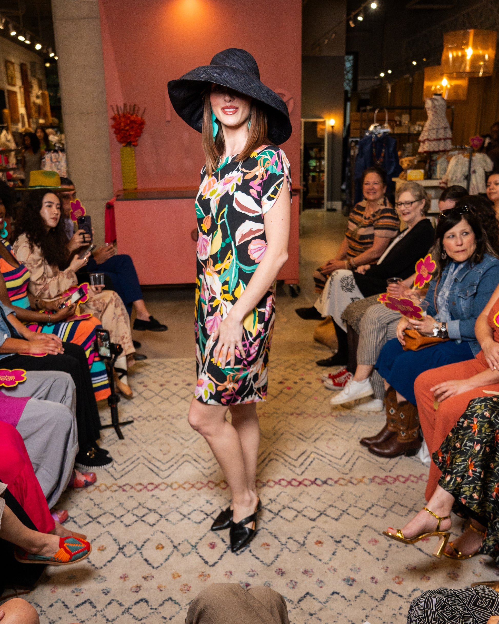 Woman in a floral dress and large hat standing in front of an audience at a fashion event.