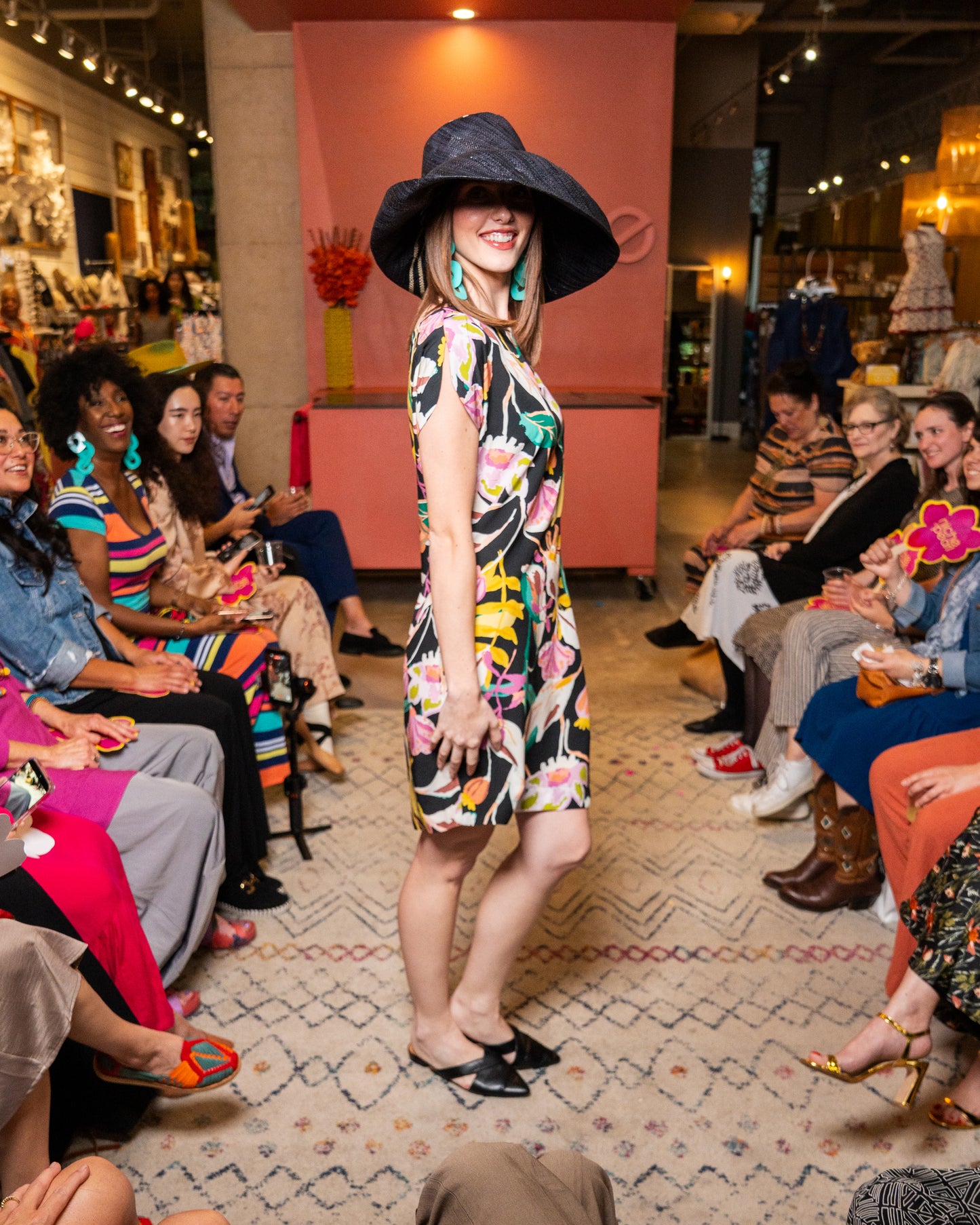 Woman in a floral dress and wide-brimmed hat walking on a catwalk with an audience watching.