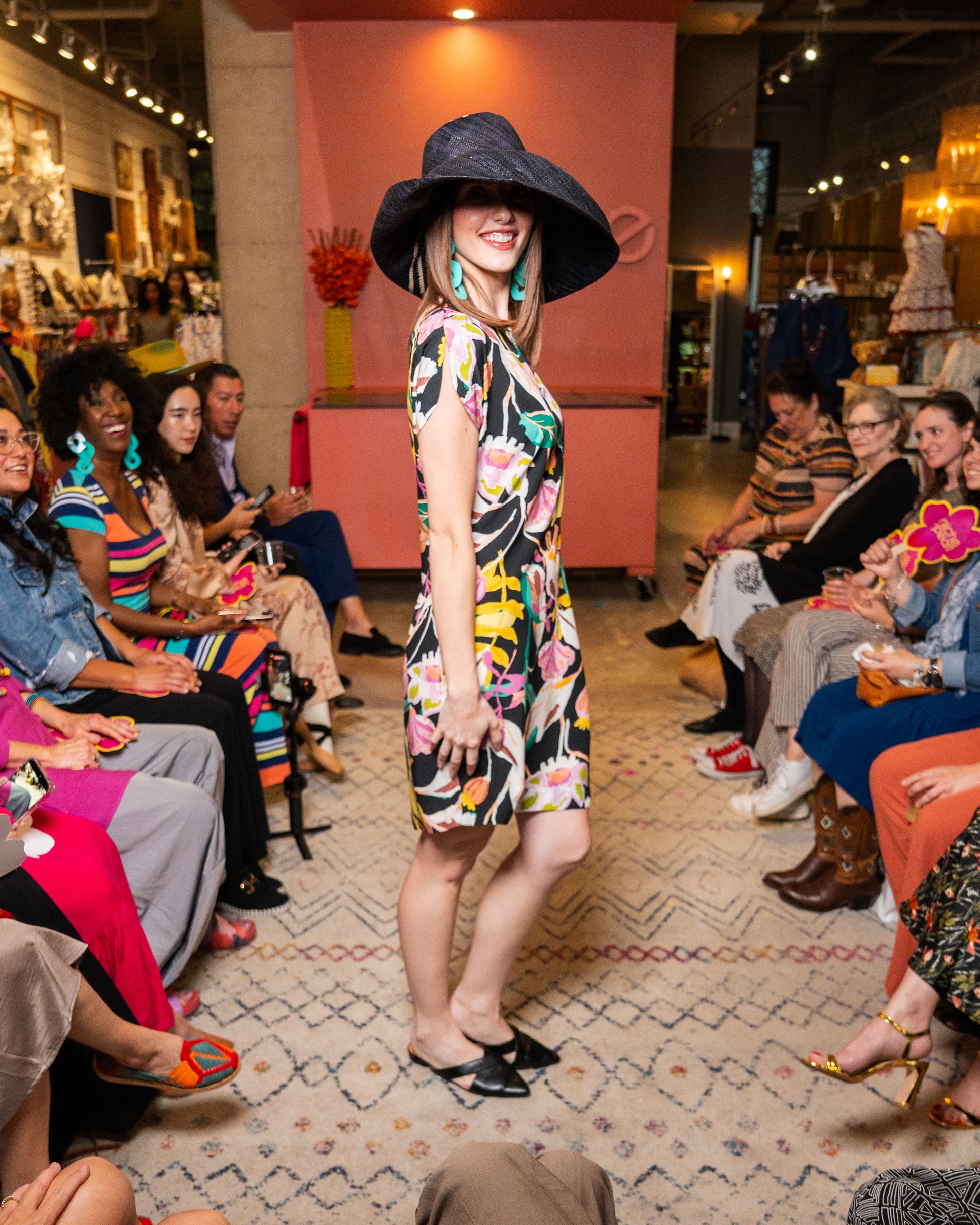 Woman in a floral dress and wide-brimmed hat walking on a catwalk with an audience watching.