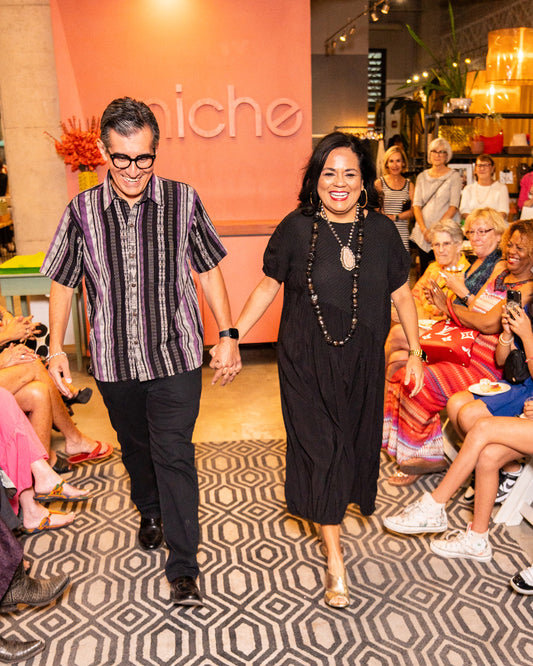 male model in a black pant with a purple, black and white striped short sleeve button down, holding hands with a female model in a black dress with a mixture of fabrics and textures