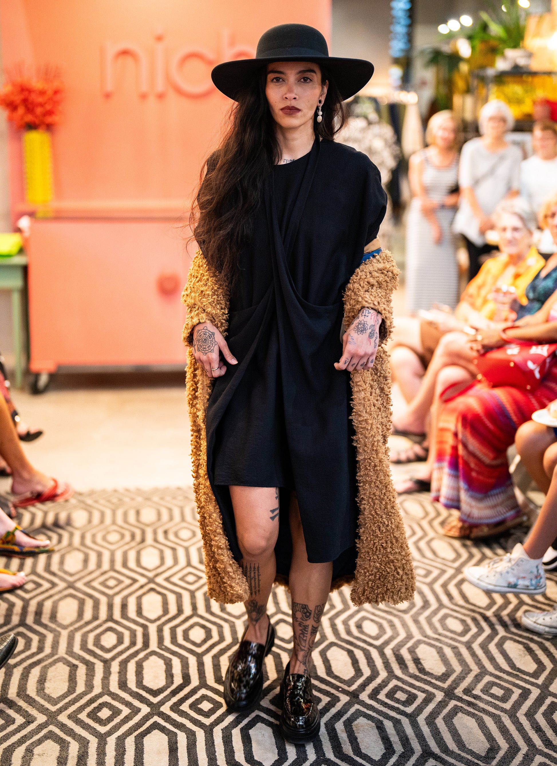 model in a brown faux fur jacket with a black dress with an adjustable strap around the neck, coming from the hips