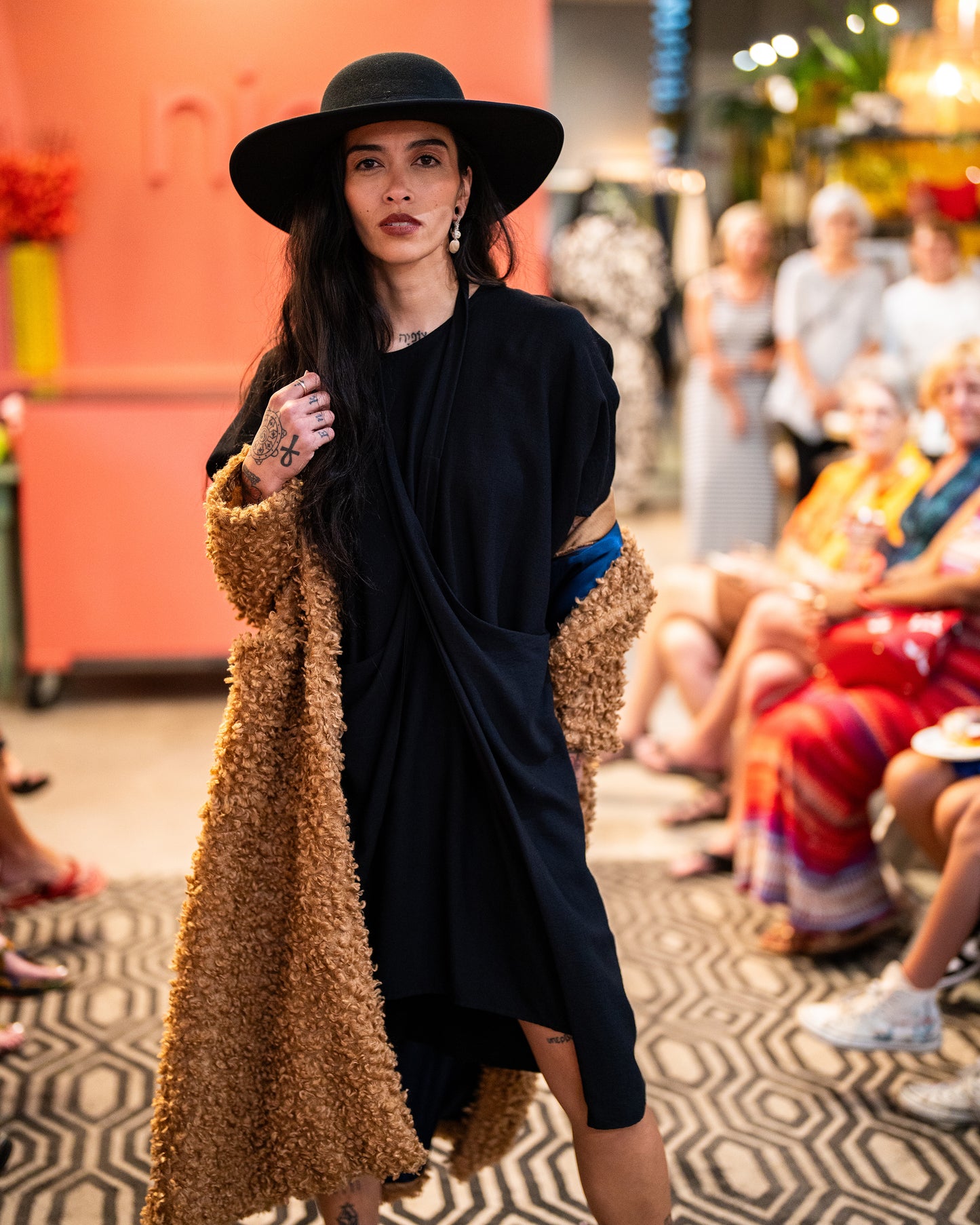 model in a brown faux fur jacket with a black dress with an adjustable strap around the neck, coming from the hips