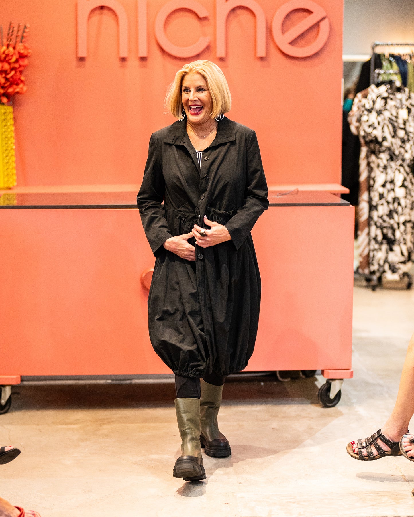 model in a black long jacket with a gathered hem, worn open over a black and white striped top, black leggings and boots