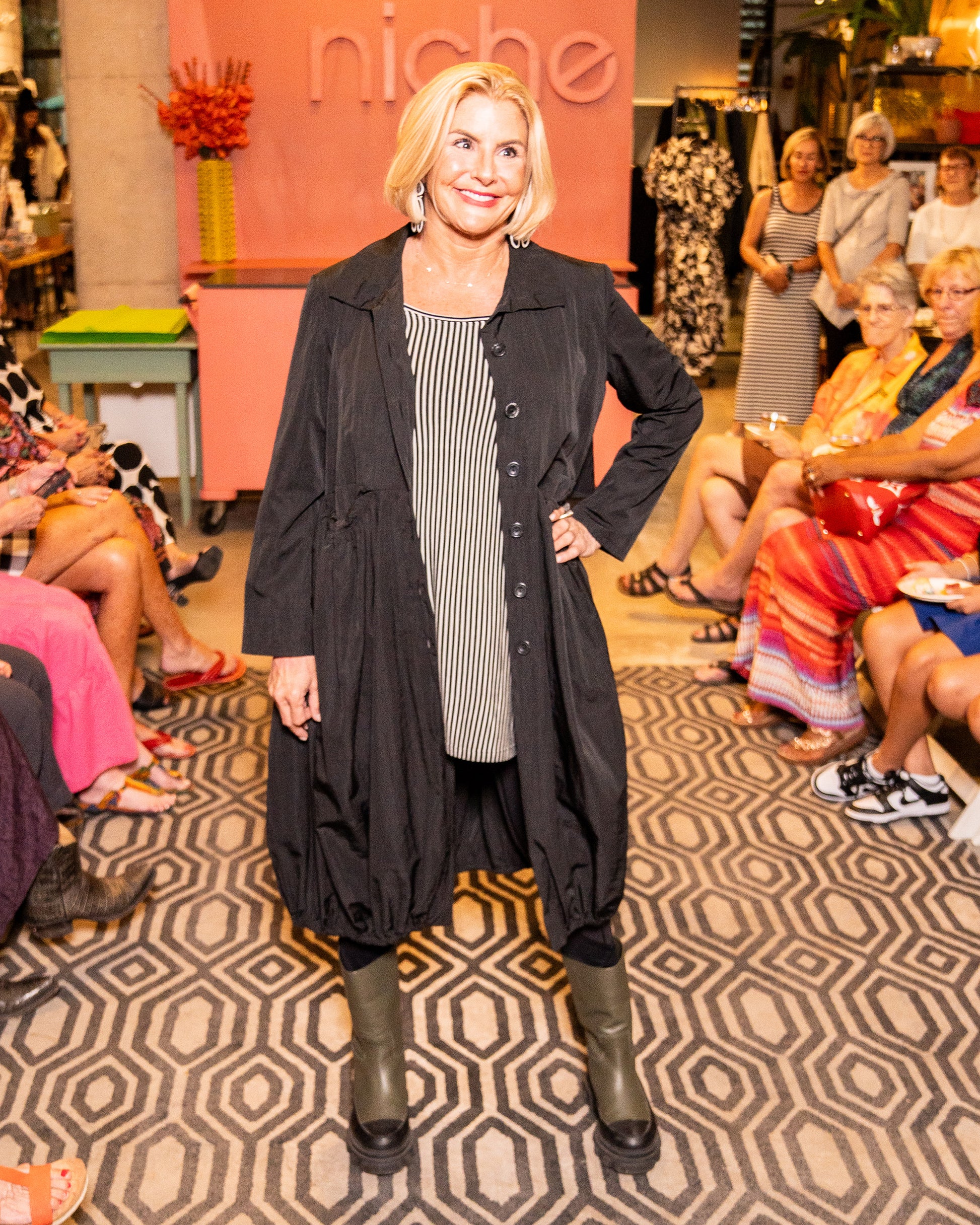 model in a black long jacket with a gathered hem, worn open over a black and white striped top, black leggings and boots