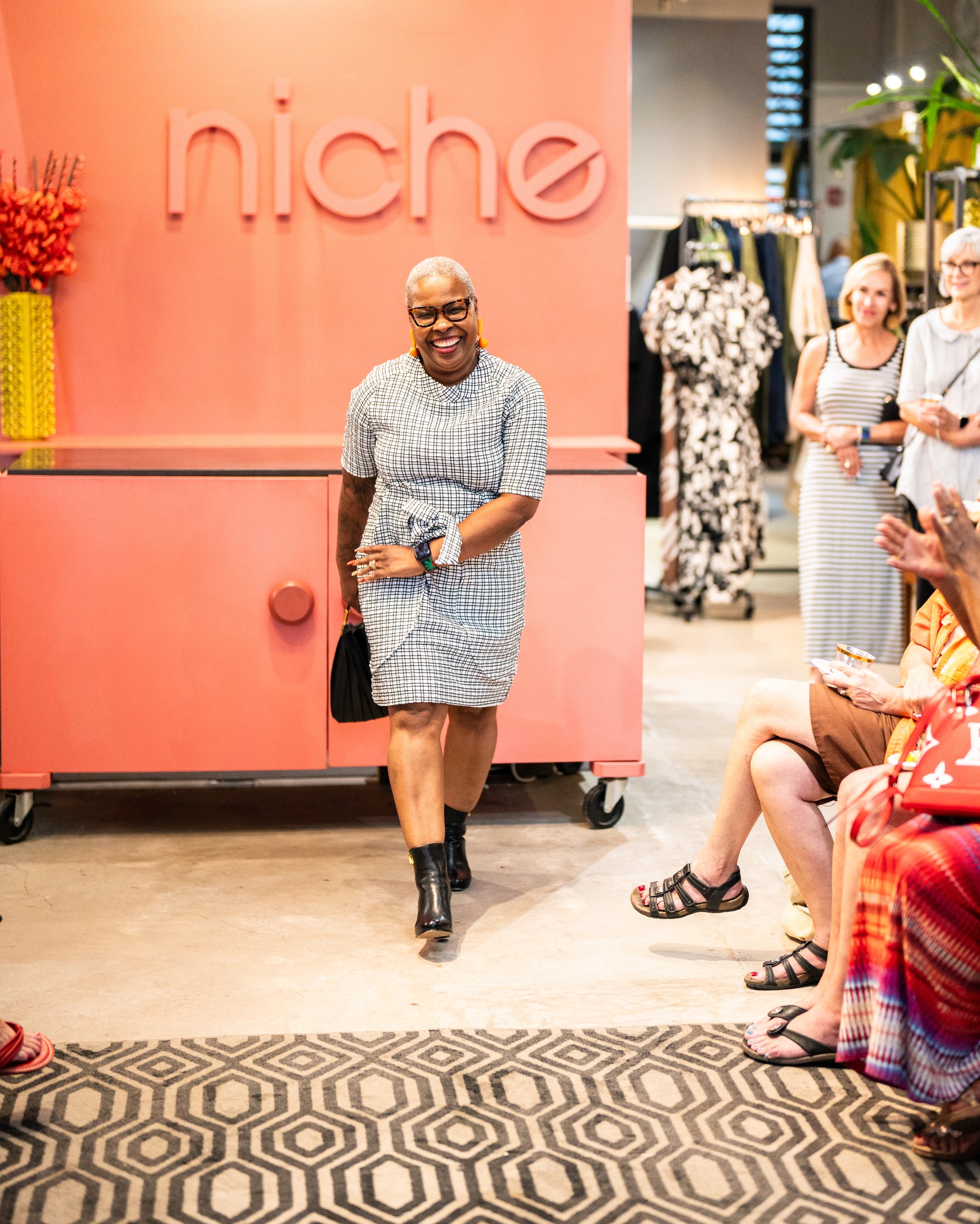 model is posing for people wearing a black and white check print raglan sleeve shift dress with a matching tie belt at the waist with black boots and a purse.