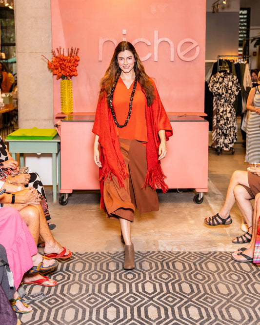 model walking down runway wearing orange v-neck top with wide leg nutmeg colored pants that have an overlapping layer in the front, burnt red cardigan and brown shoes