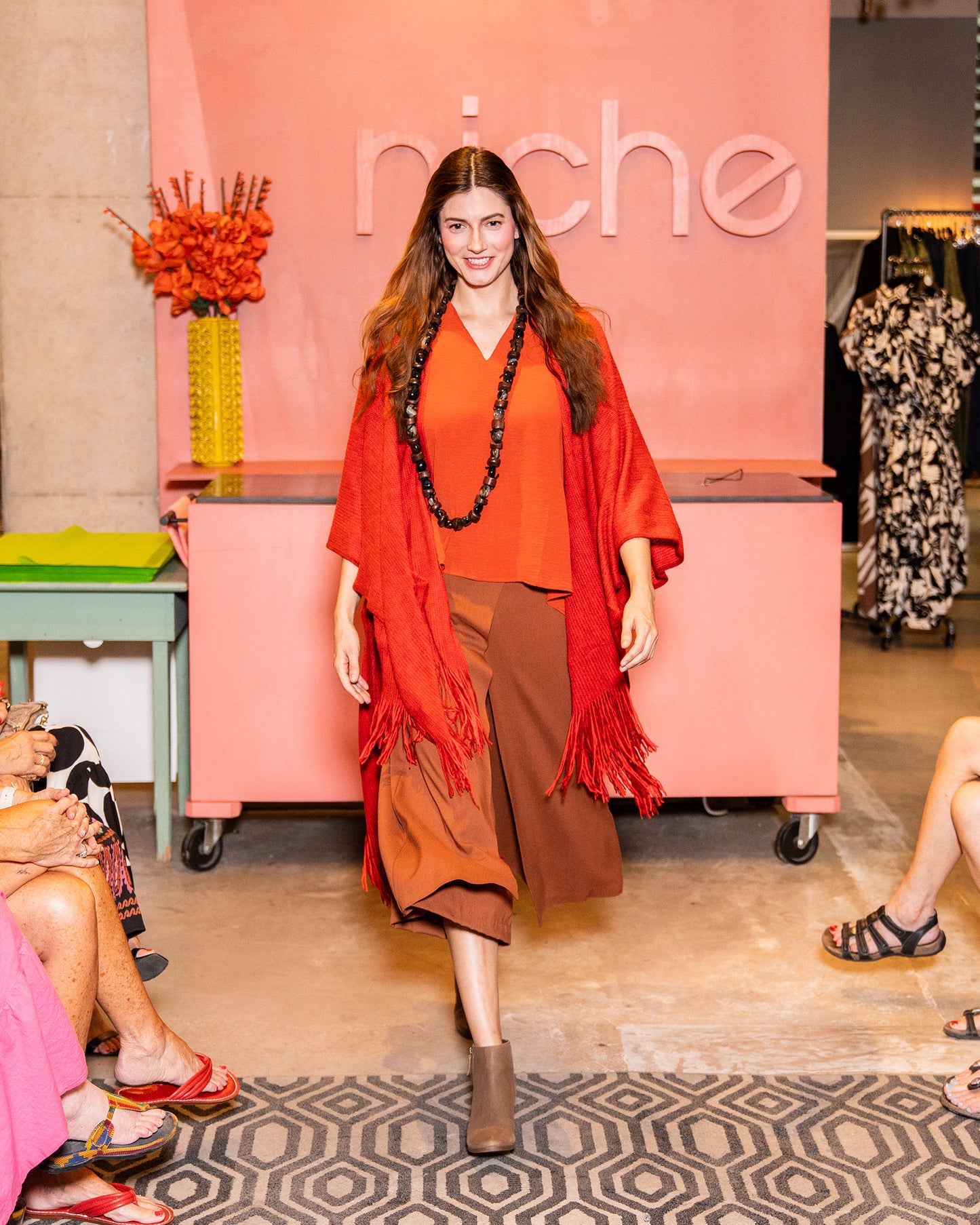 model in a rust color wide leg pant with an orange shirt and fire red shawl with a fringe hem