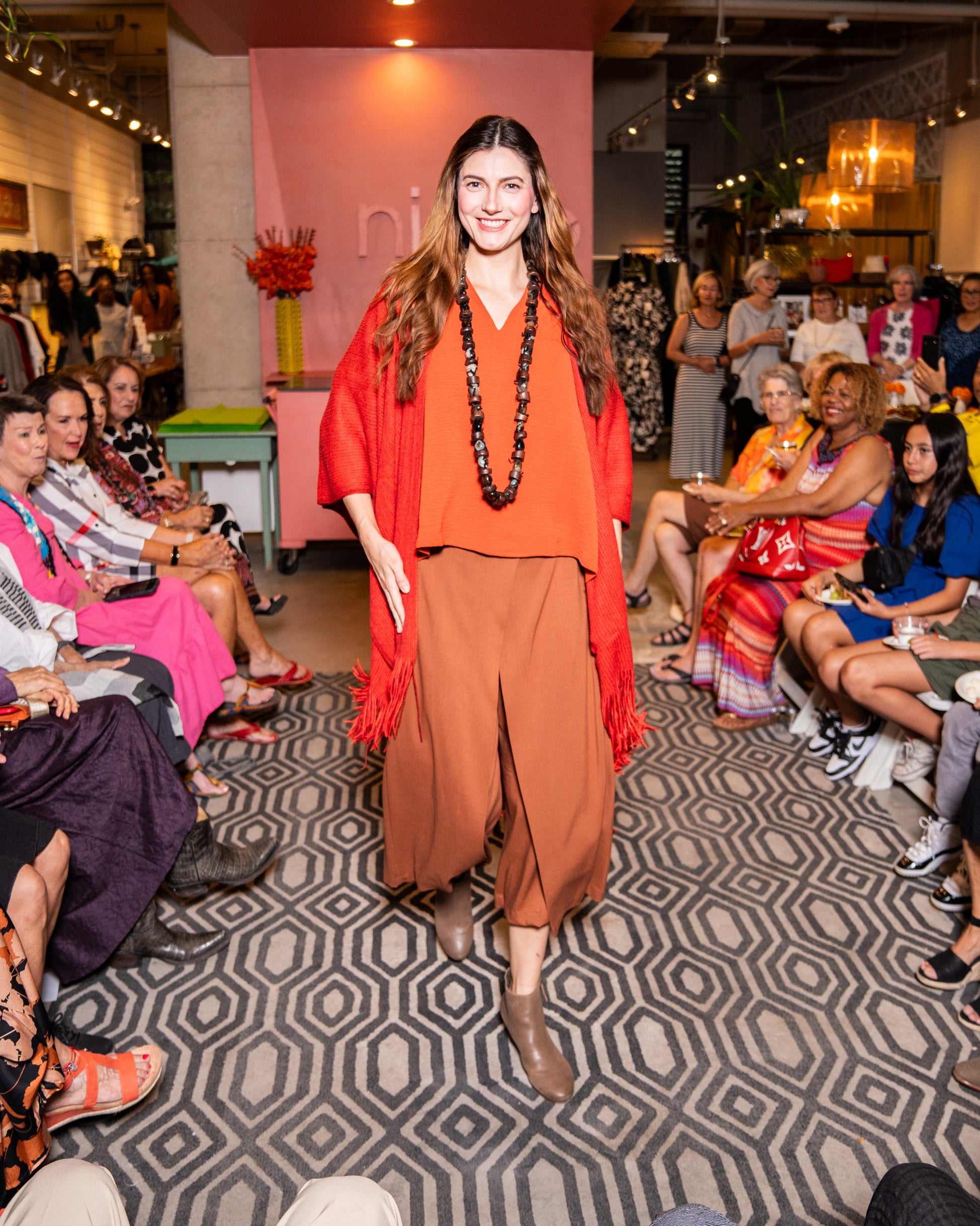 model walking down runway wearing orange v-neck top with wide leg nutmeg colored pants that have an overlapping layer in the front, burnt red cardigan and brown shoes