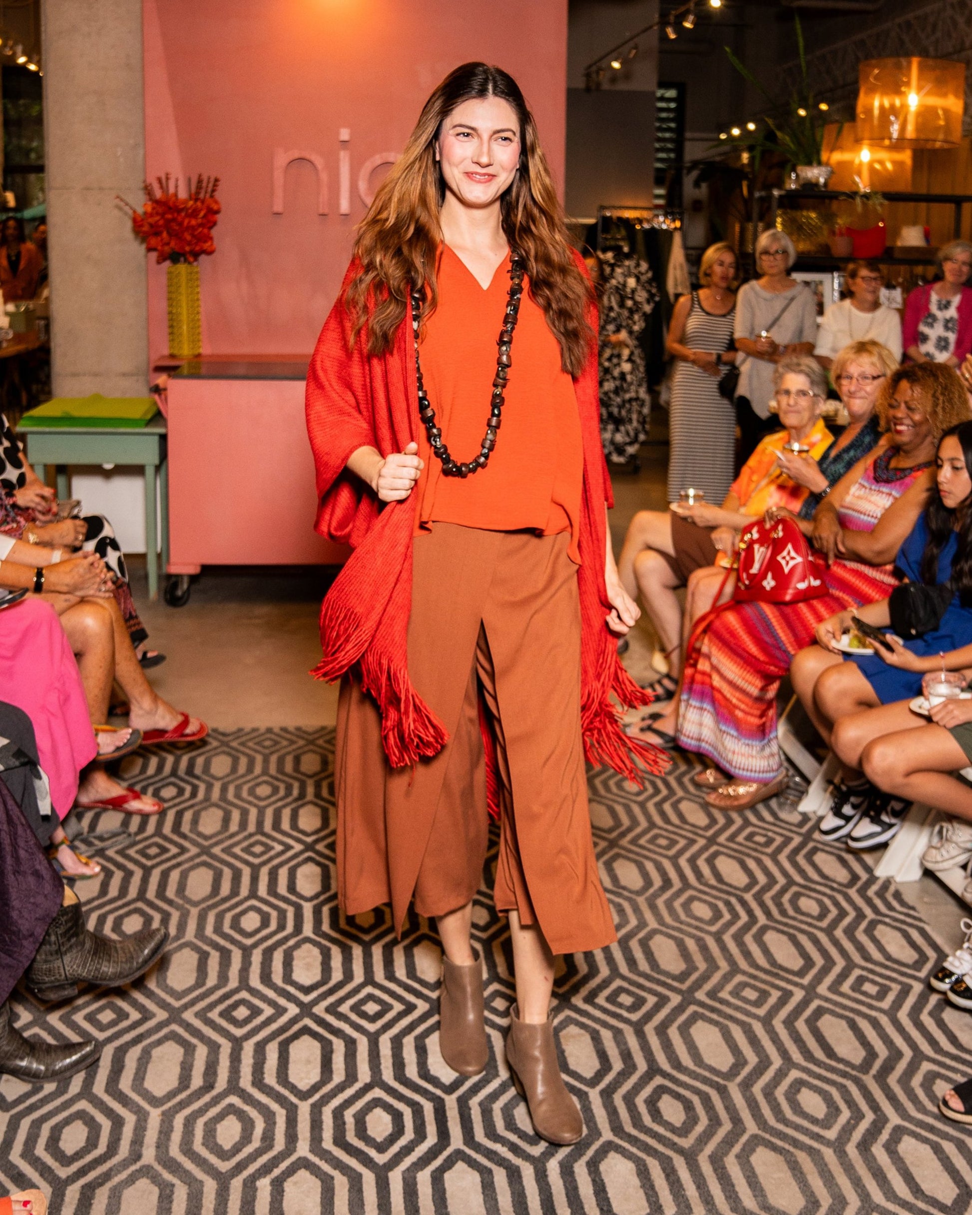 model in a rust color wide leg pant with an orange shirt and fire red shawl with a fringe hem