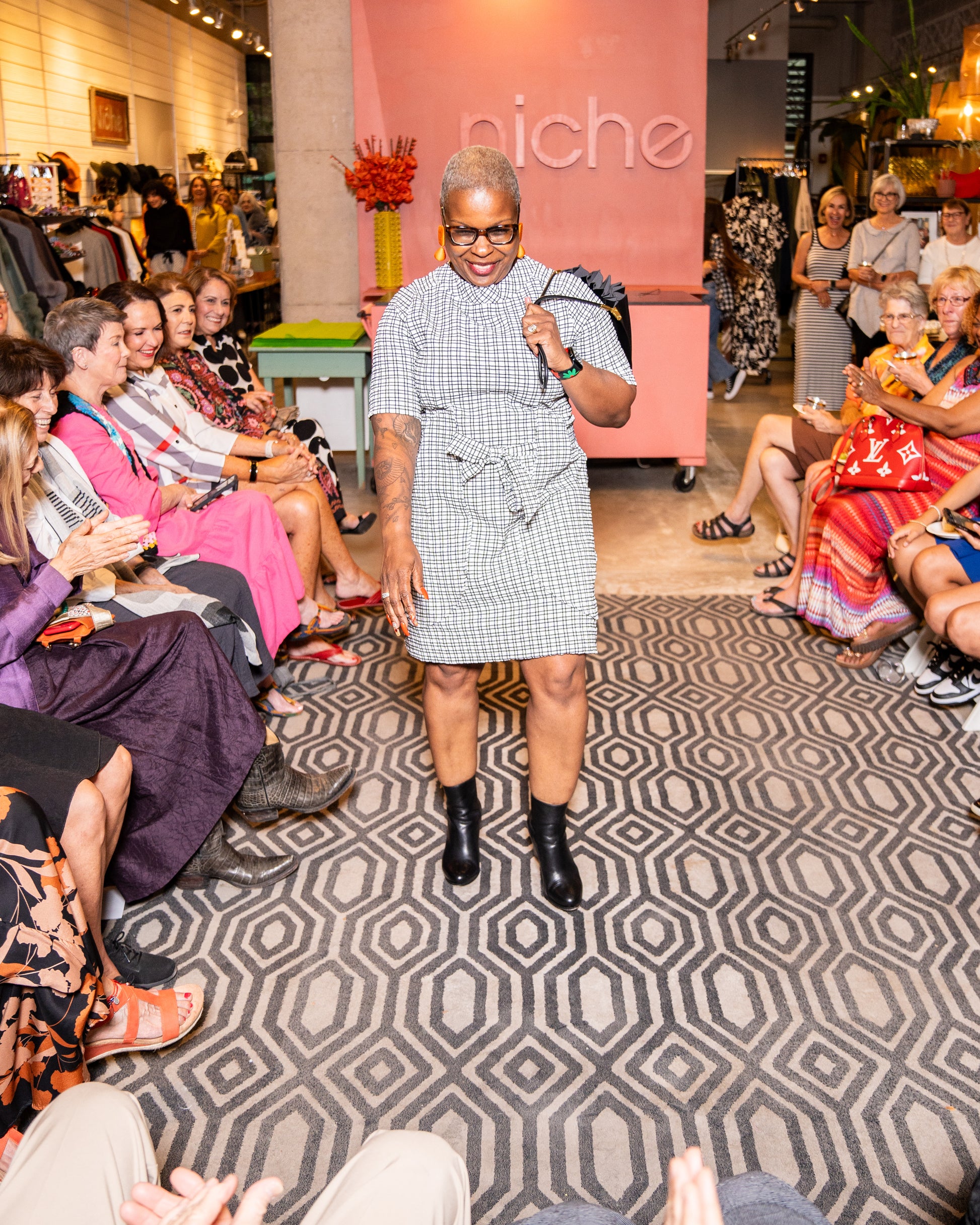 model is posing for people wearing a black and white check print raglan sleeve shift dress with a matching tie belt at the waist with black boots and a purse.