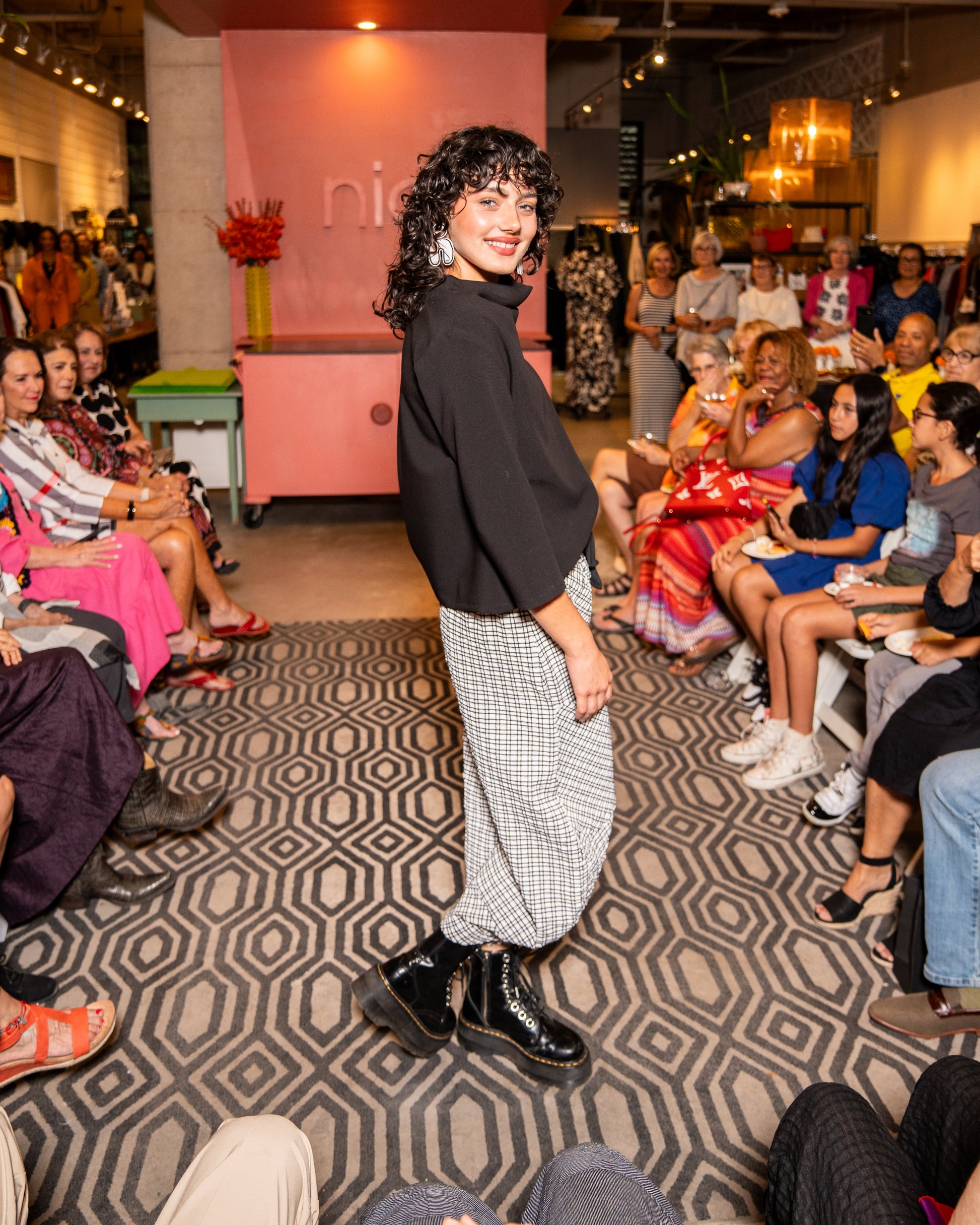 model walking down runway wearing black cowl top with 3/4 sleeves and black and white check pants with black tie belt