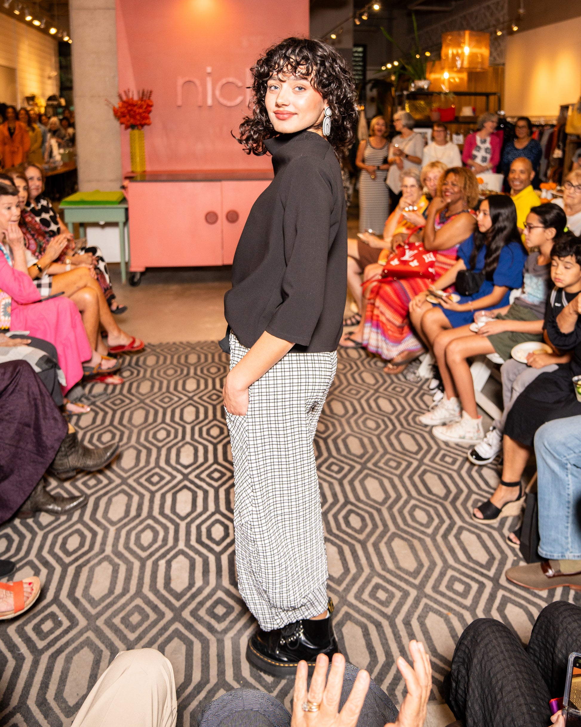 model walking down runway wearing black cowl top with 3/4 sleeves and black and white check pants with black tie belt