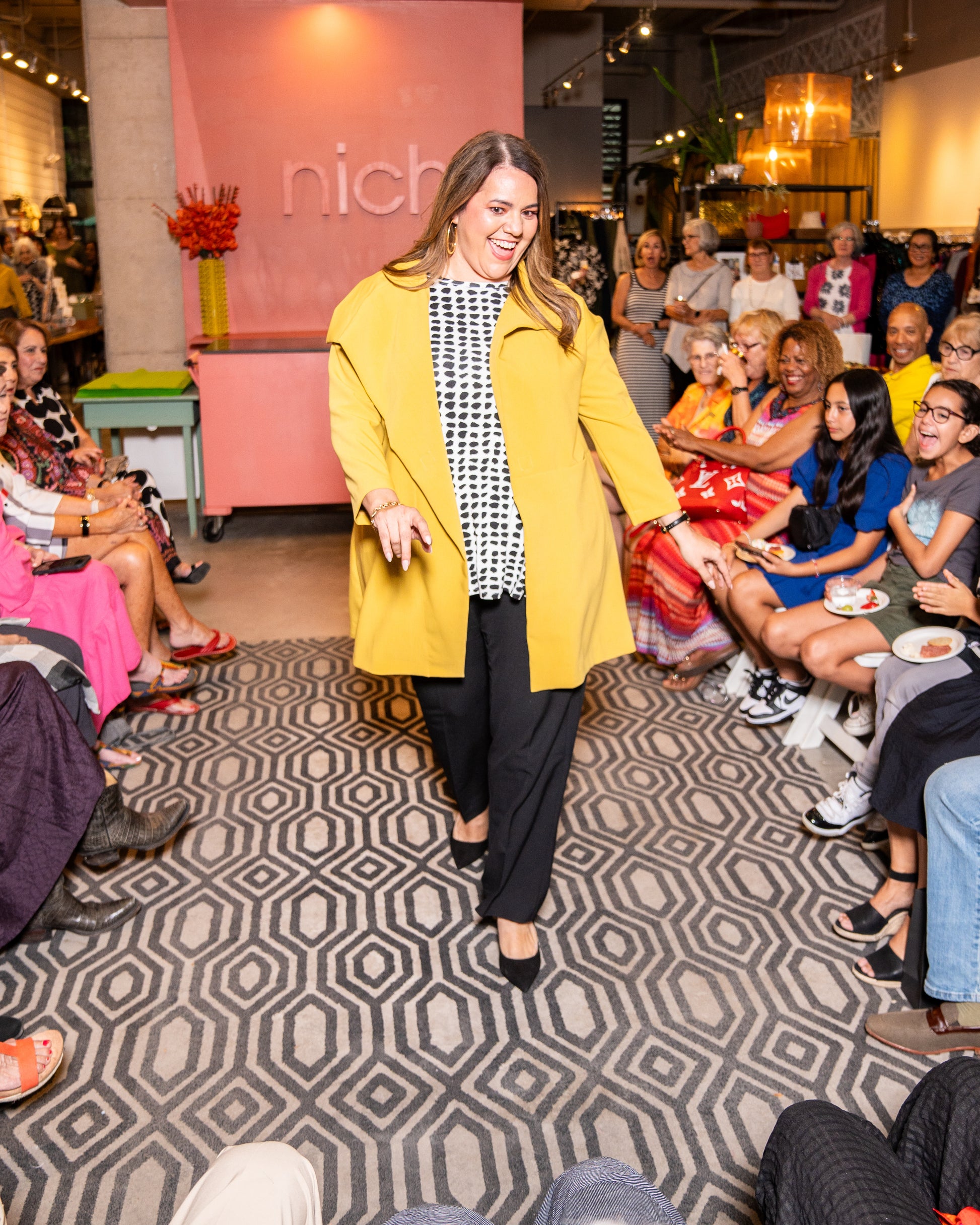 model posing for people wearing black pants with a gold asymmetrical jacket with a large collar and black and white polka dot top underneath.