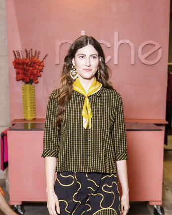 Model walking on a catwalk at a fashion event with wearing yellow check button up top, swirl pattern pants and yellow bandana